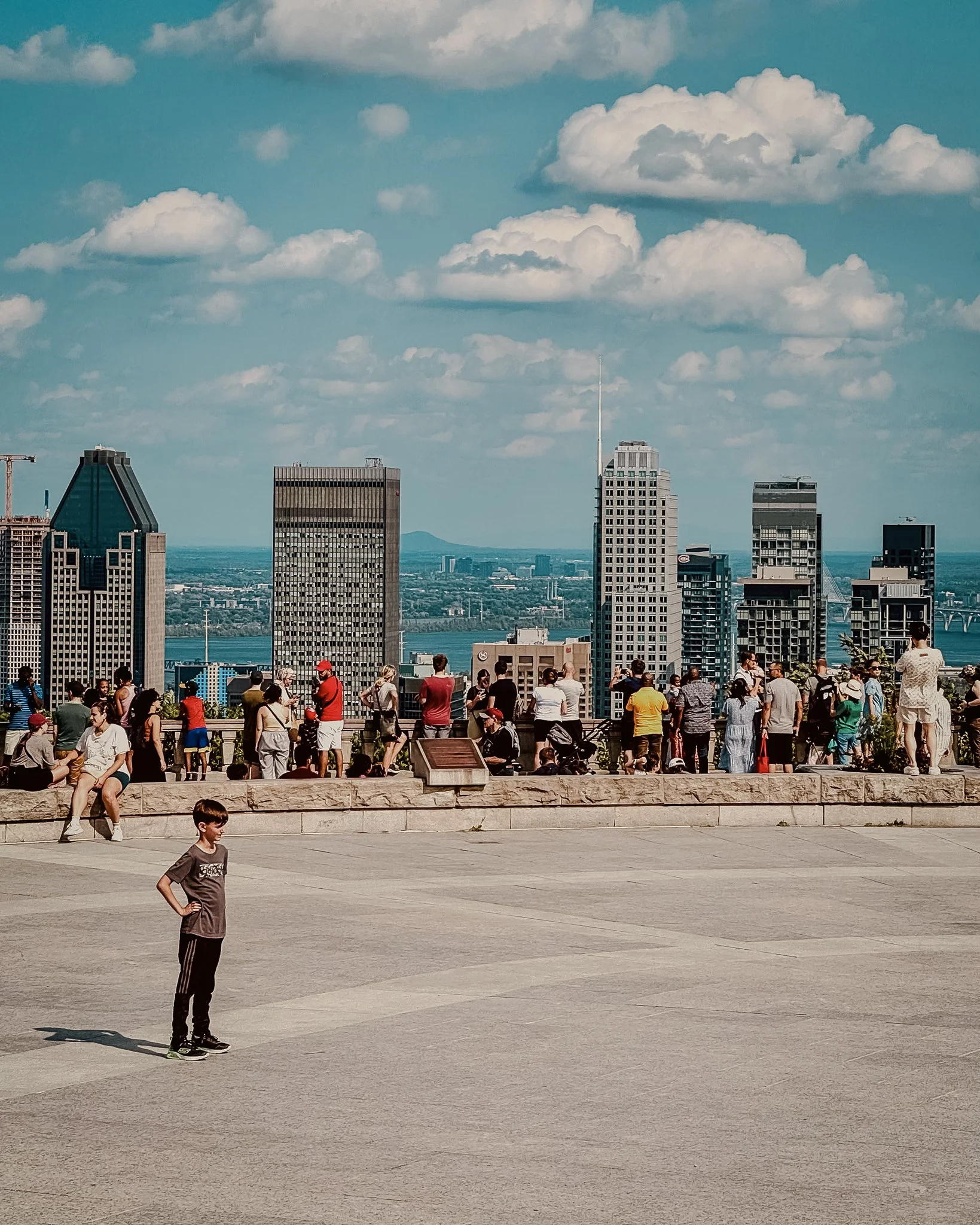 A young boy standing on a wide open concrete space with a city skyline and a river in the background, under a partly cloudy sky.