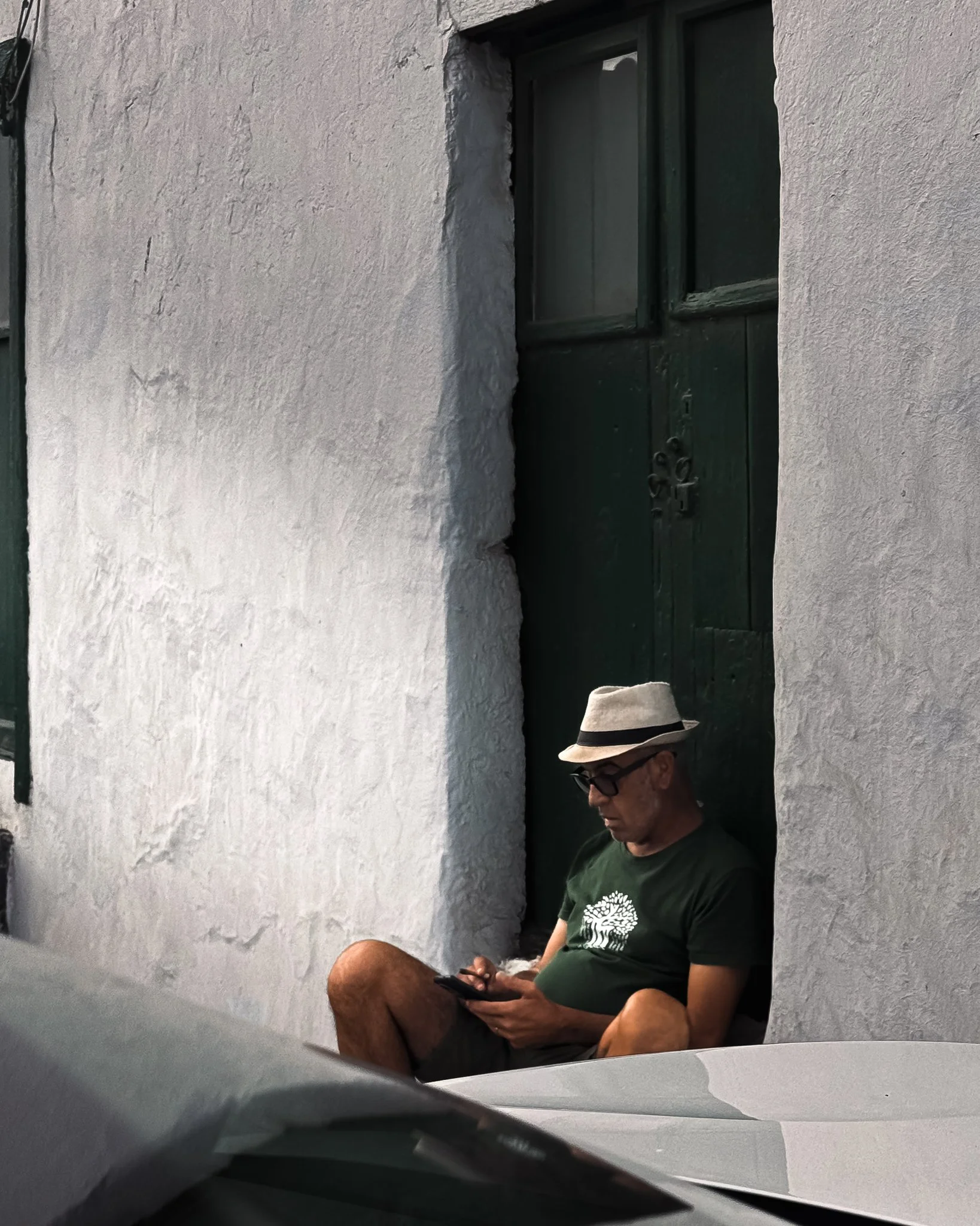 A man sitting against a white stucco wall, wearing sunglasses, a light-colored fedora, a dark green t-shirt with a tree graphic, and shorts, looking at his phone.