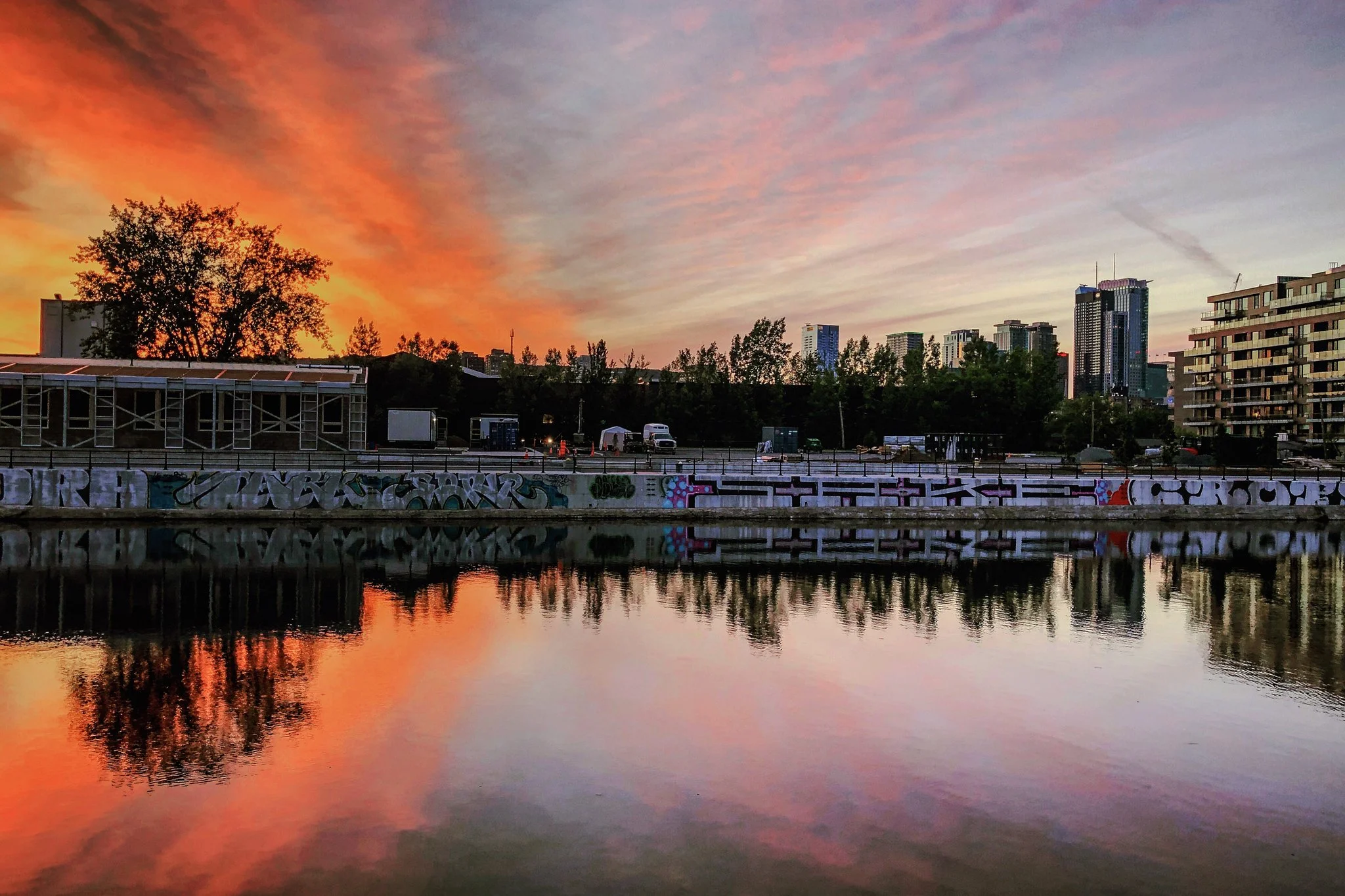 Sunset over an urban river with city buildings in the background, graffiti on a concrete barrier, and reflection in the water.