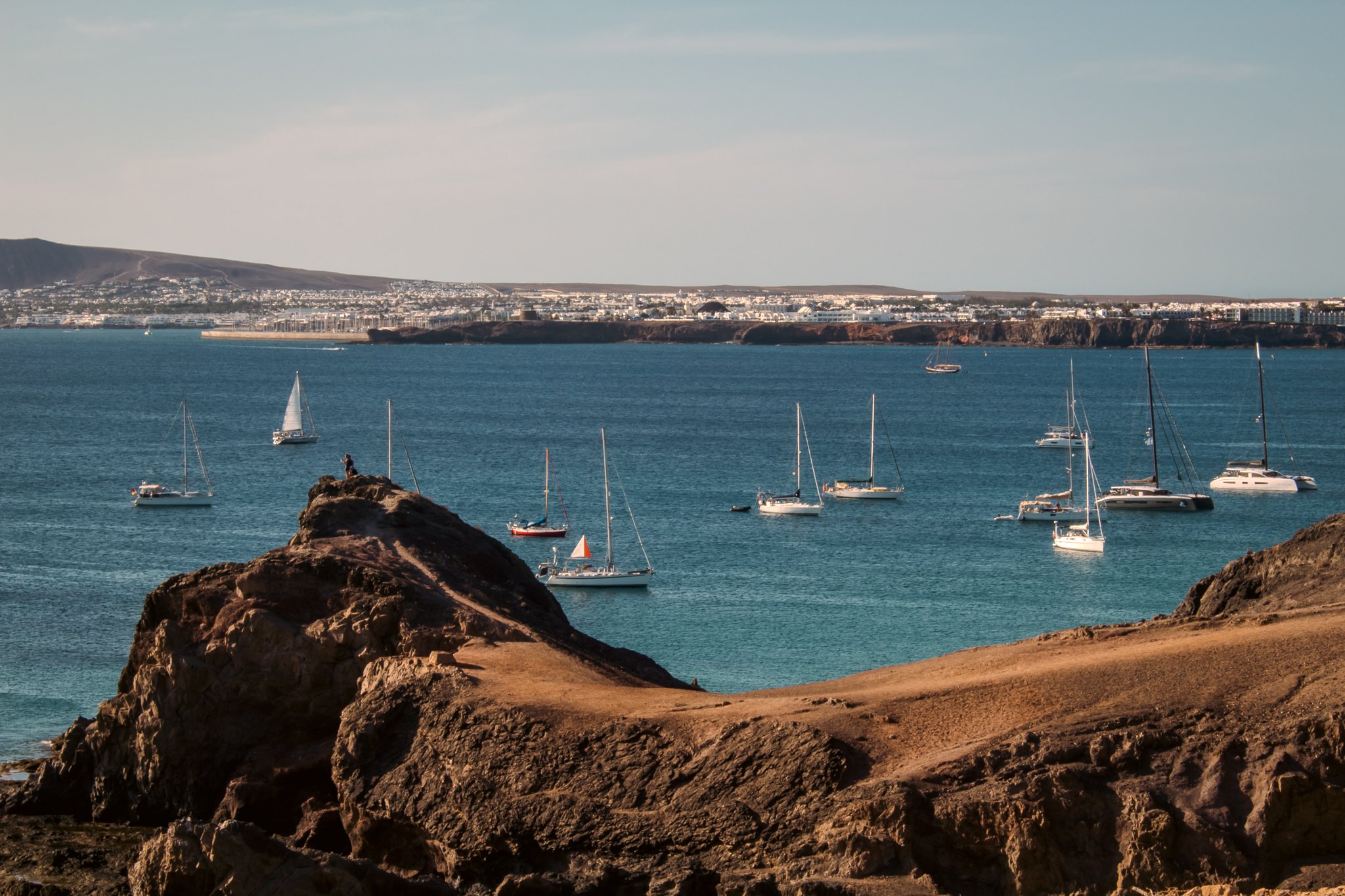 A coastal landscape with rocky foreground, a blue sea with sailboats, and a distant shoreline with buildings and hills under a partly cloudy sky.