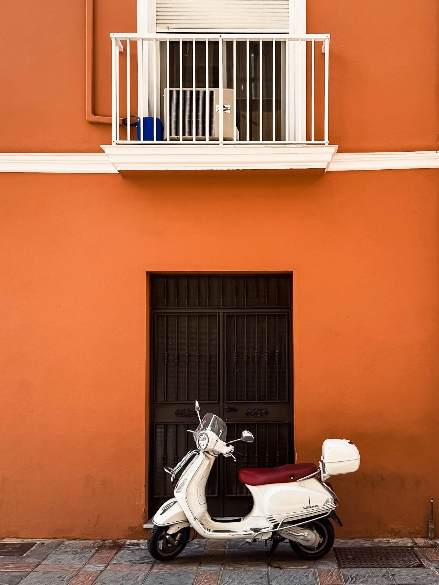A white scooter with a red seat and a white storage box parked in front of an orange building with a black door and a balcony with a white railing.