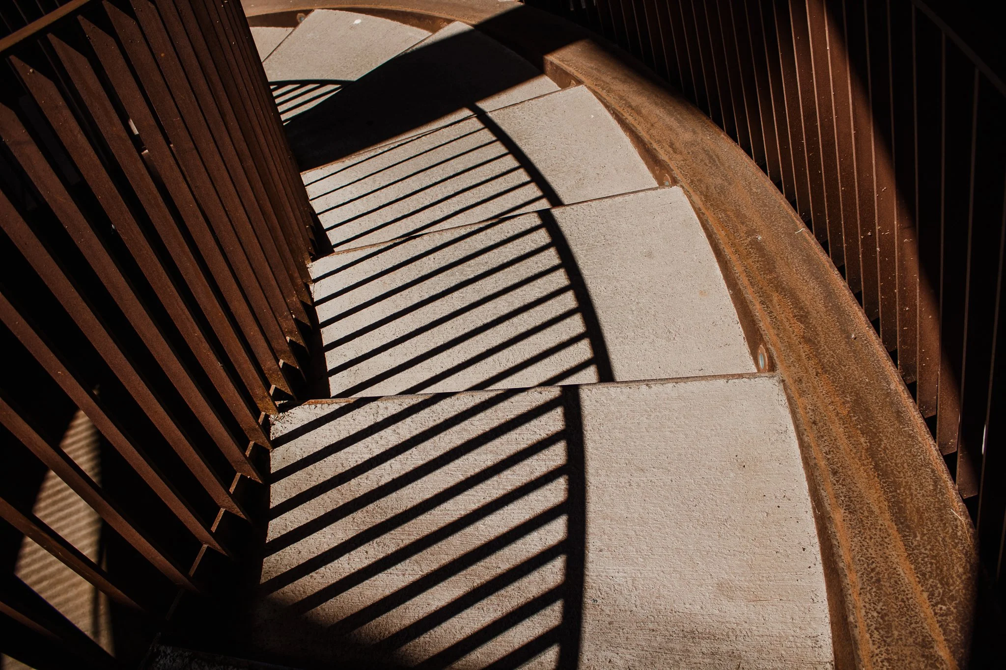 Curved outdoor staircase with concrete steps and rust-colored metal railings casting striped shadows.