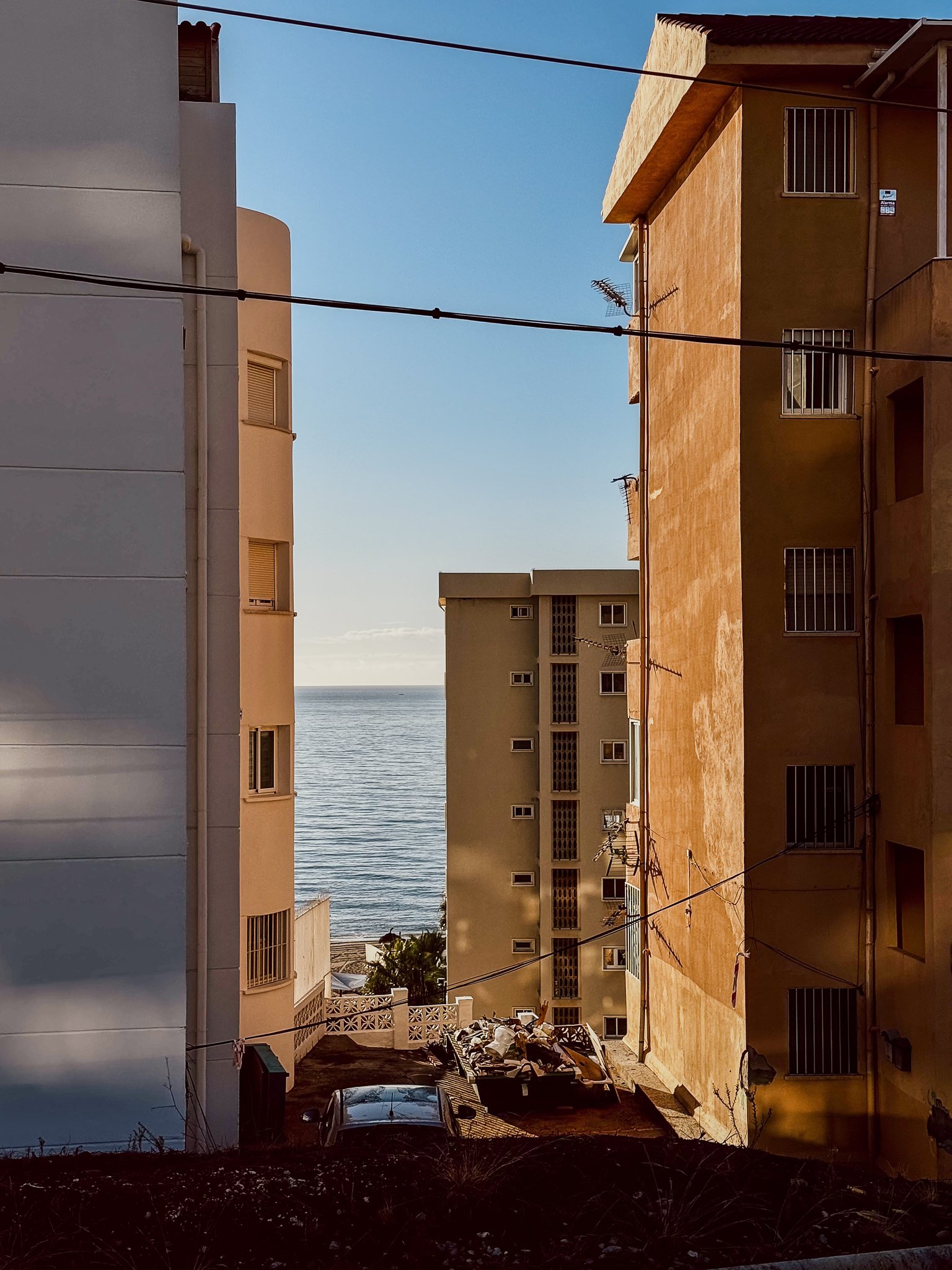 View of a narrow alley between buildings leading to the ocean in the background. The buildings are multi-story with balconies and windows, painted in white, beige, and yellow. There are electrical wires crisscrossing above, and trash and a car are at the bottom of the alley.