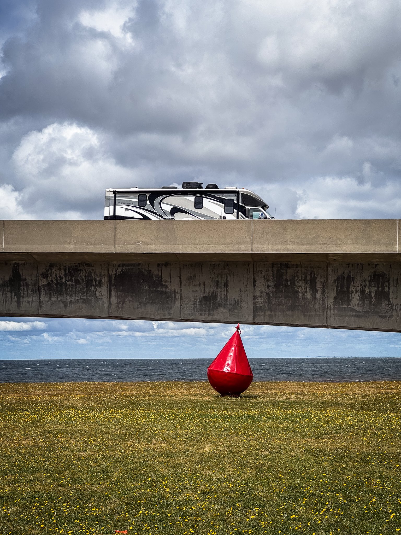A recreational vehicle (RV) parked on top of a bridge over a grassy area near the sea, with a large red buoy in the foreground and dark clouds in the sky.