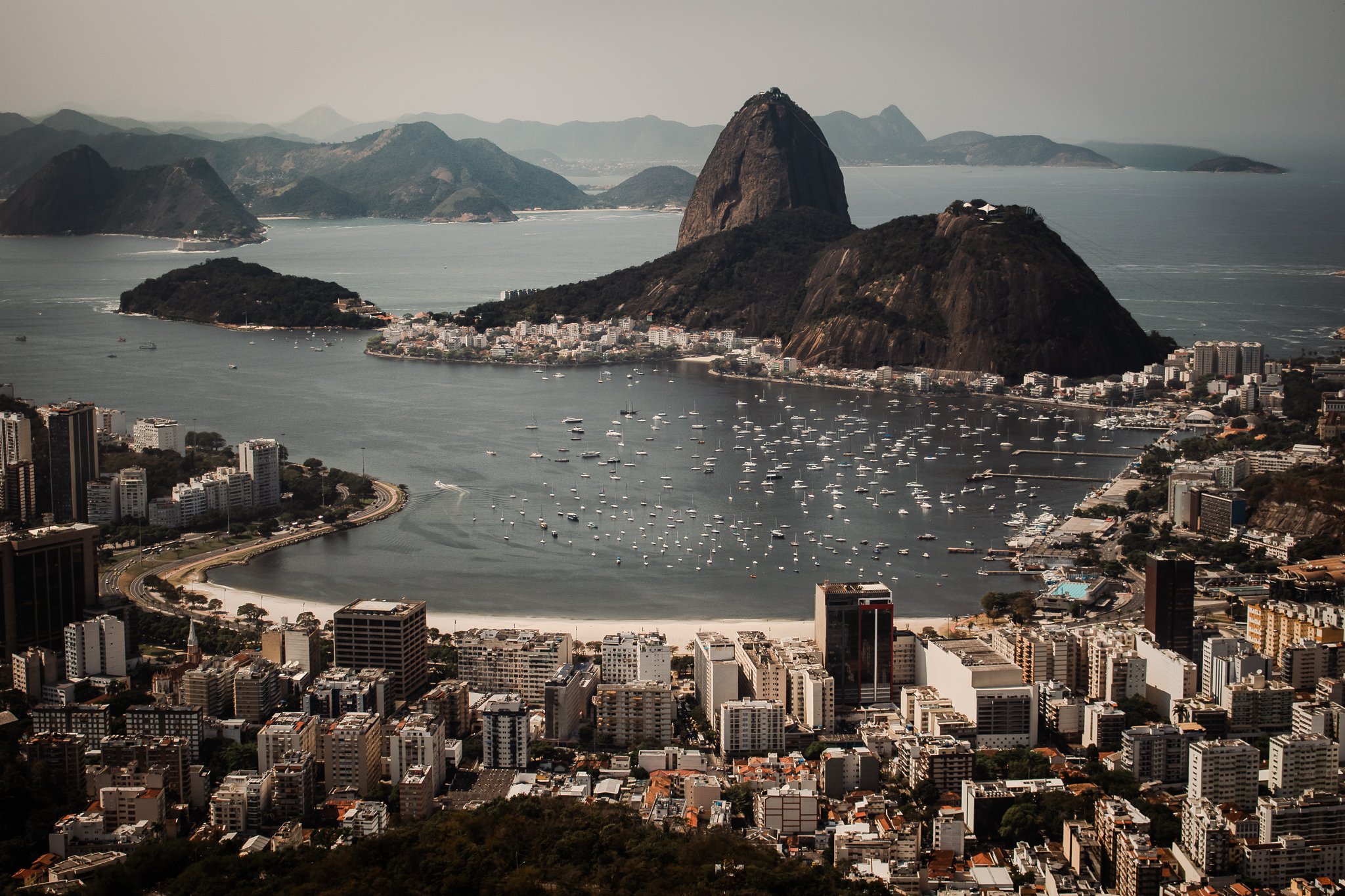 A cityscape of Rio de Janeiro with Sugarloaf Mountain in the background, overlooking the harbor filled with numerous boats and yachts, surrounded by high-rise buildings and lush hills.