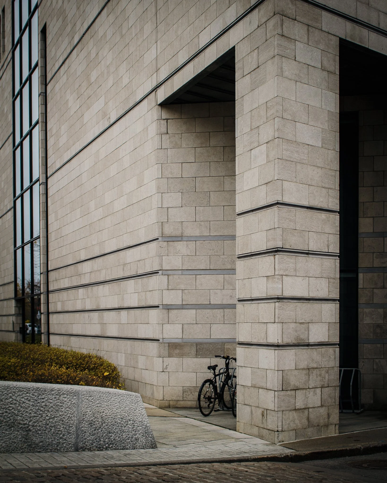 A modern building with large glass windows and beige brick walls. A bicycle leans against the corner of the building near a small hedge and a stone sculpture.