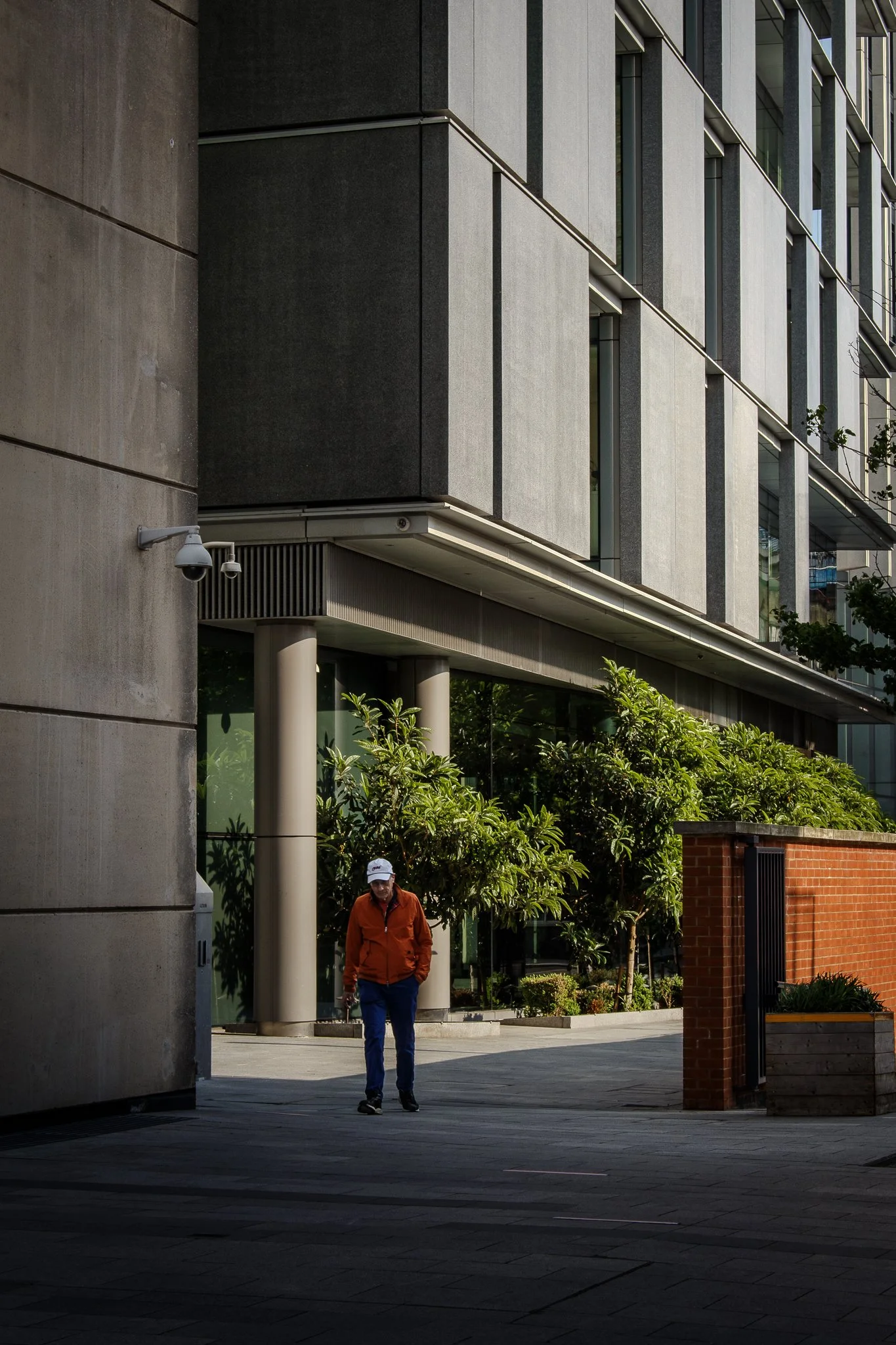 A person in an orange jacket and a white cap walking in an urban area surrounded by modern buildings and greenery.