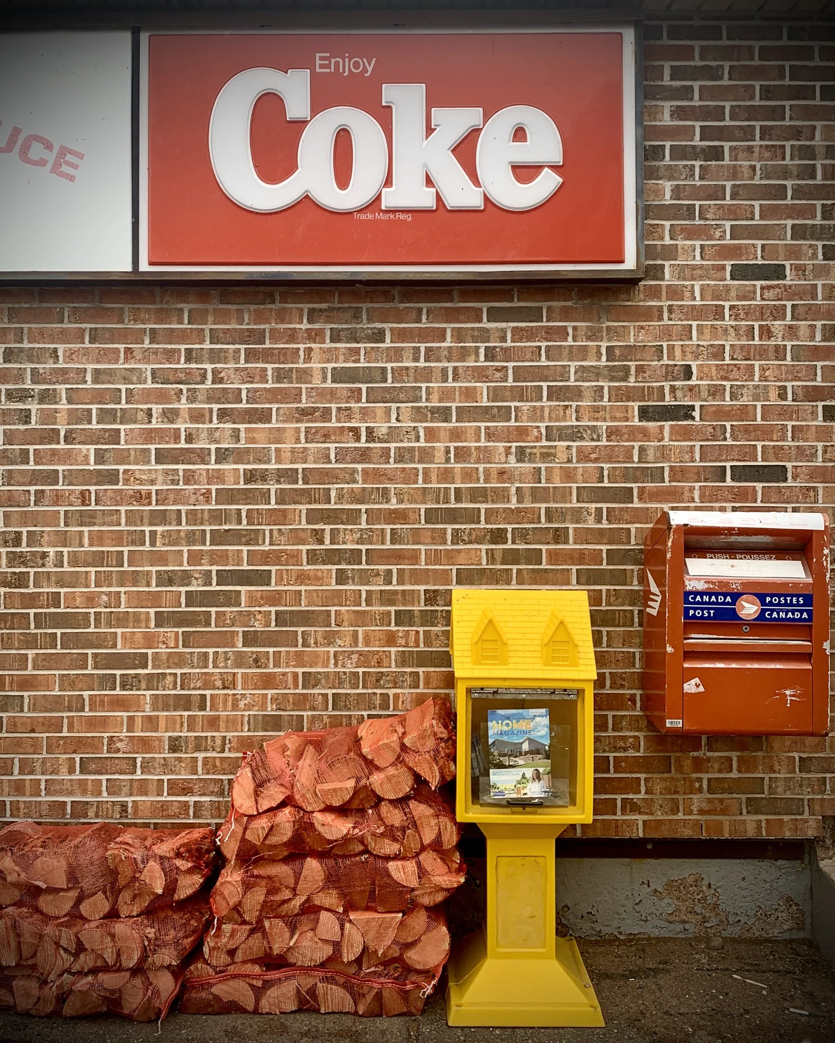 A brick wall with a red sign that reads 'Enjoy Coke' at the top. To the right, there is a red Canada Post mailbox. In front of the wall, there is a yellow newspaper box with a house-shaped top, containing magazines, and a pile of firewood next to it.