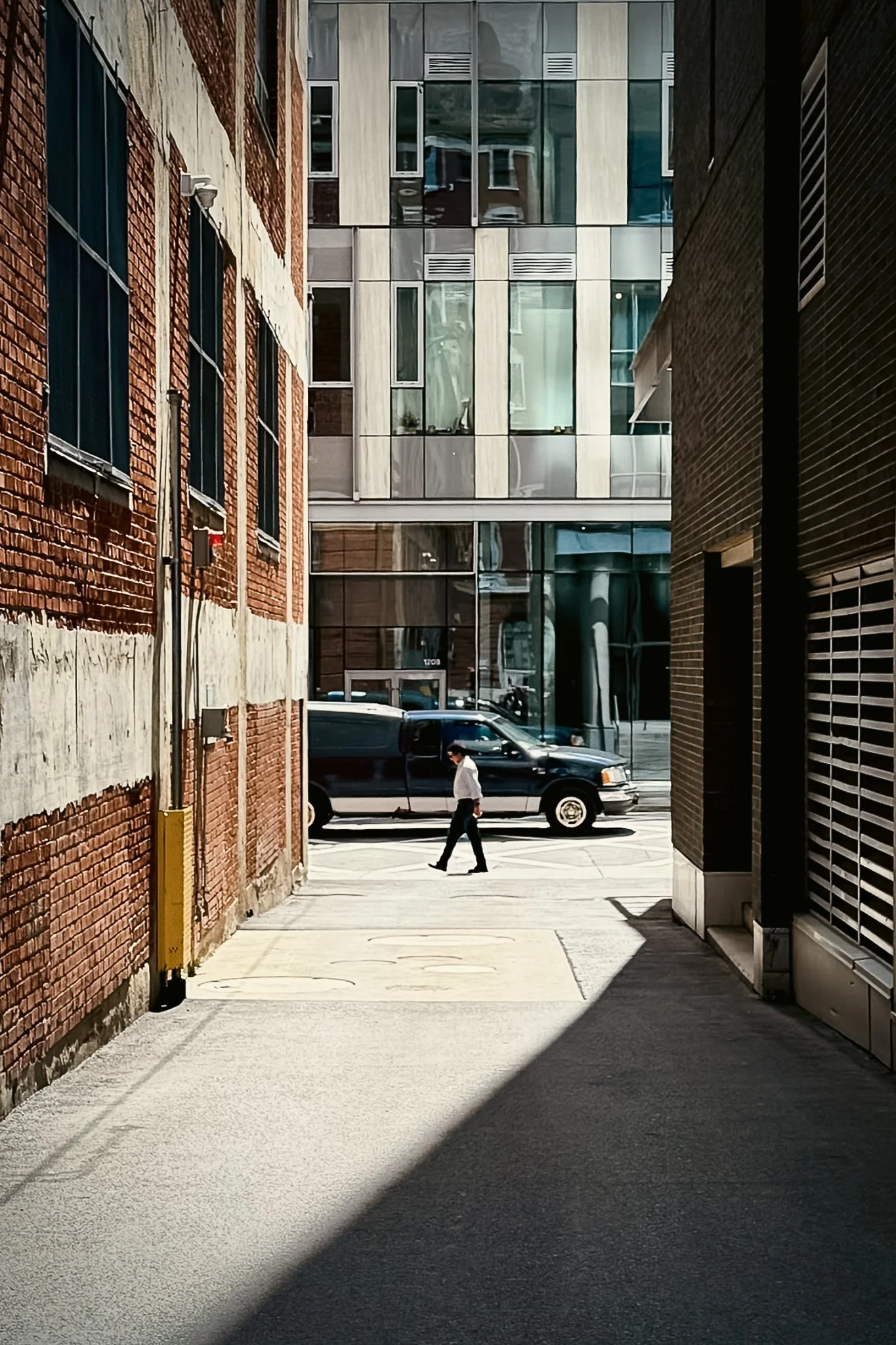 A man walking across a city alleyway with a black vehicle parked at the curb and modern glass buildings in the background.