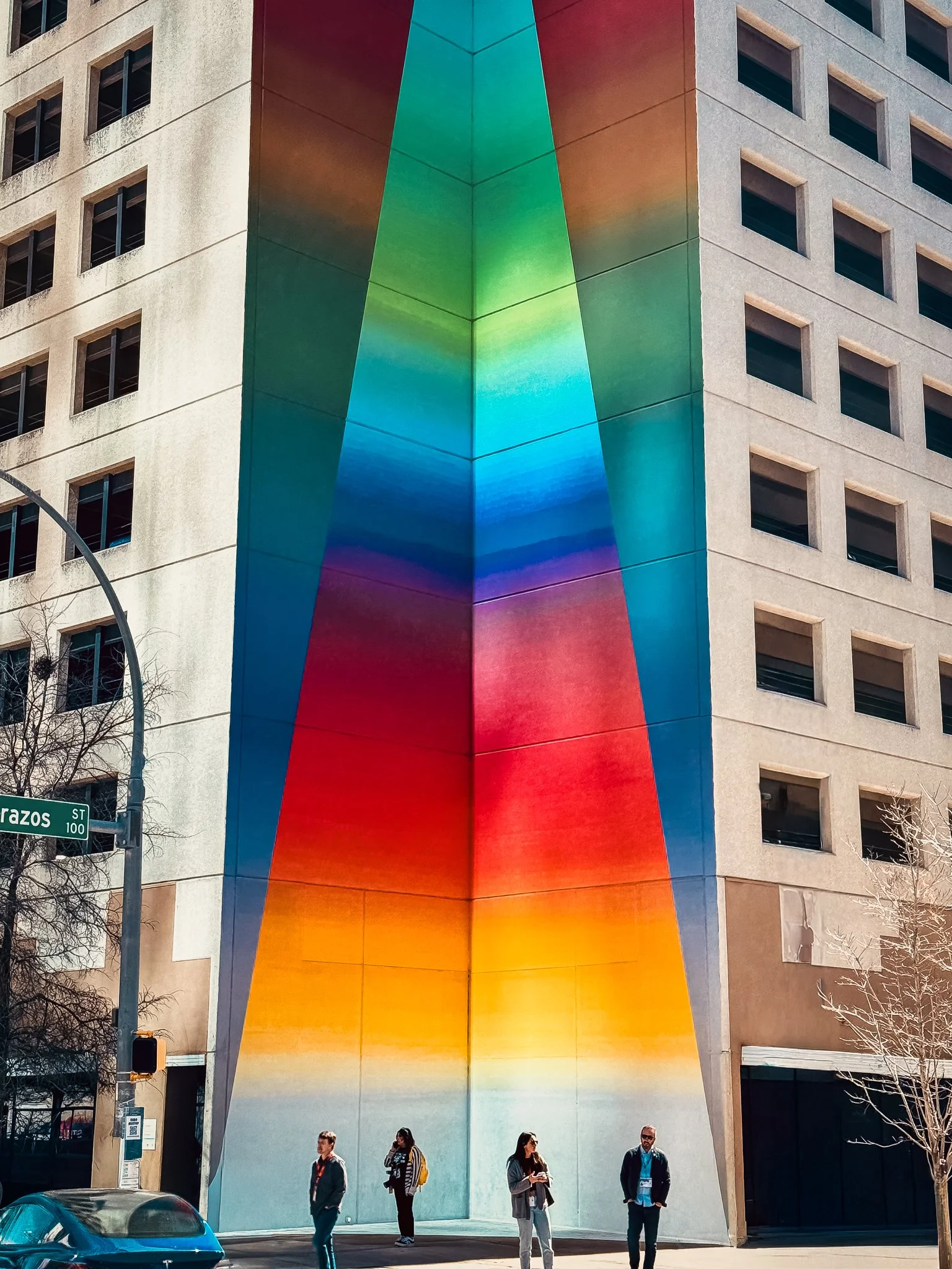 Colorful, rainbow-colored mural on the corner of a building in an urban setting, with several pedestrians and a street sign reading "Paz Street 100".