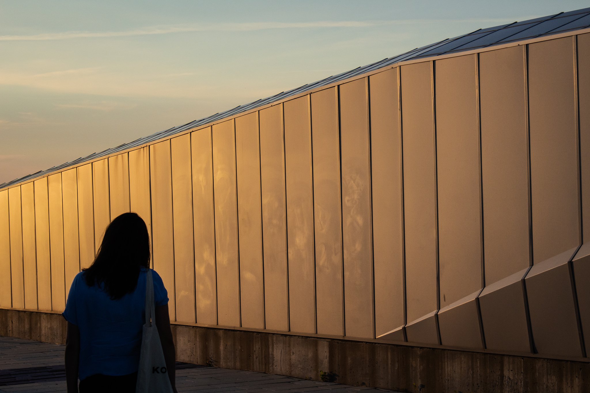 A woman with dark hair wearing a blue shirt carries a white tote bag as she walks past a modern building with a reflective gold exterior during sunset.