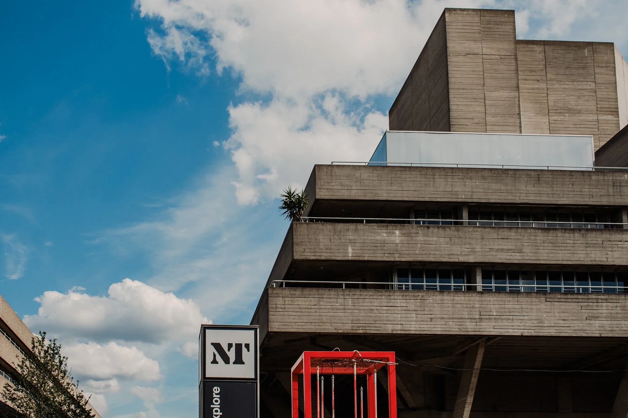 A modern concrete building with multiple levels and a small palm tree on a balcony, under a blue sky with clouds, a sign that reads 'NT' and an outdoor red swing surrounded by a frame.