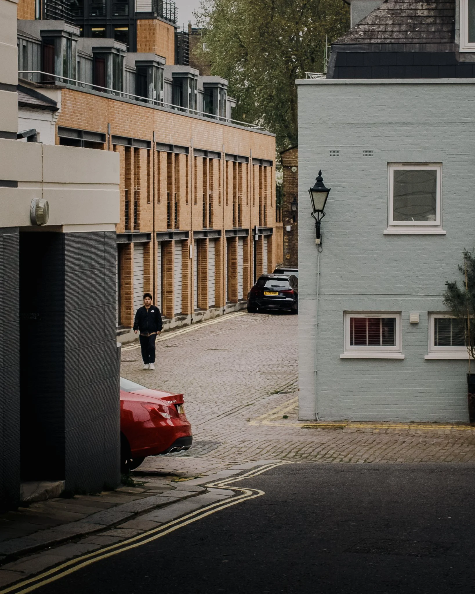 A person walking on a cobblestone street in front of a tan brick building with black window frames and a white house with a black lamp post and one window. There are parked cars, including a black one and a red one.