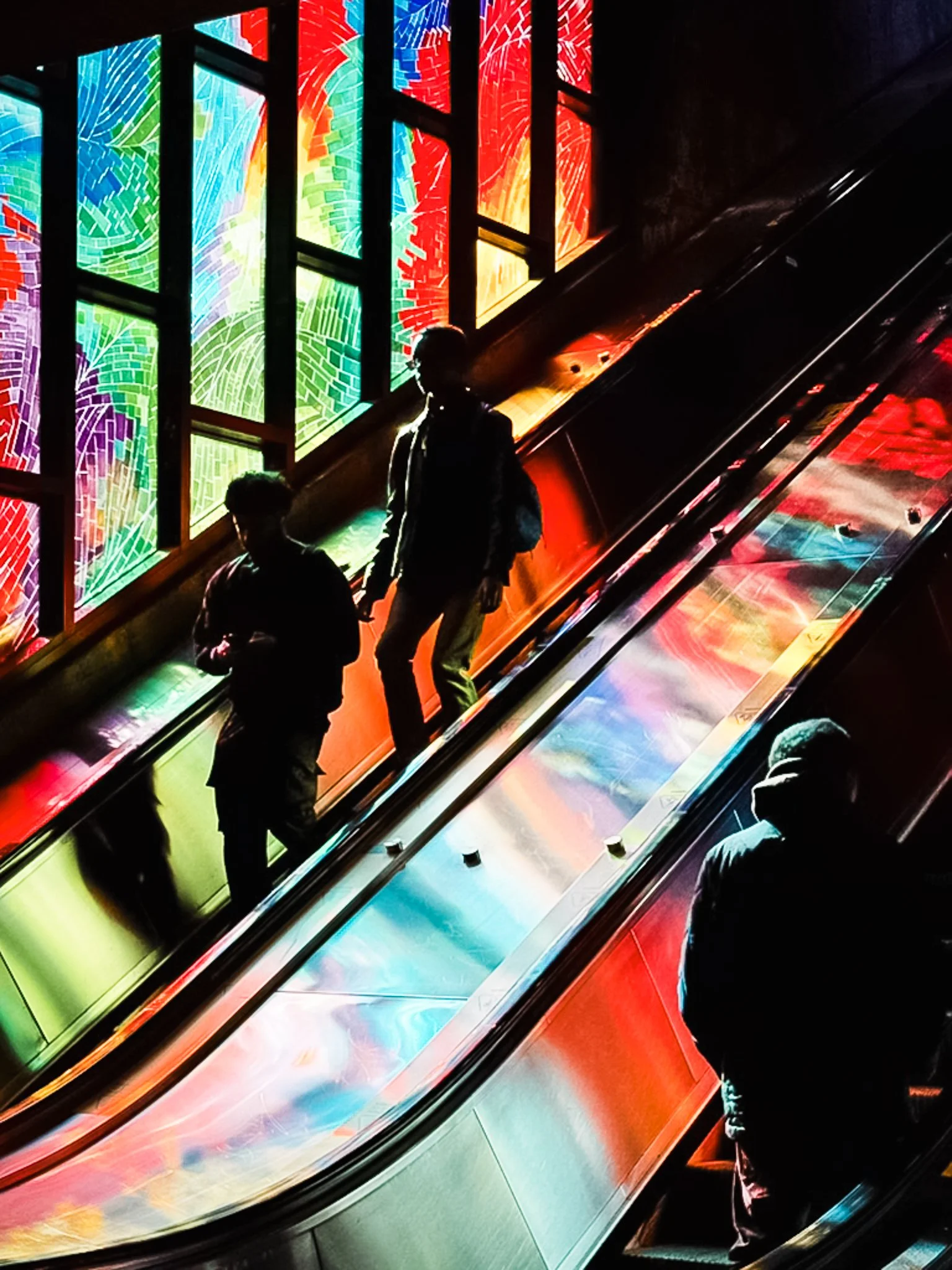 Three people walking on an escalator in front of a colorful stained glass window with vibrant, abstract patterns.