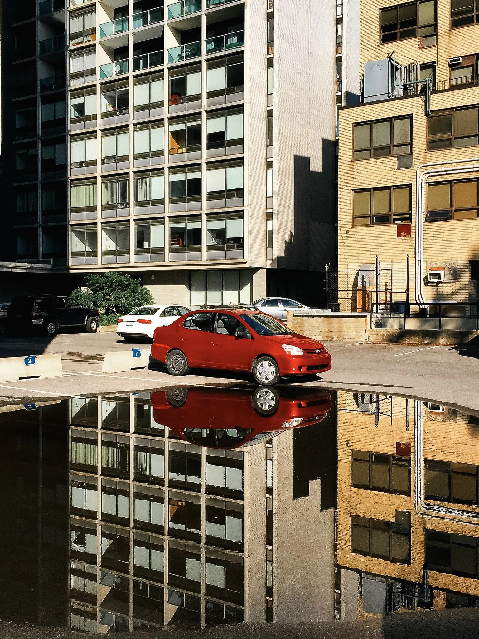 A red car parked in a parking lot with a tall building in the background. The car's reflection is visible in a puddle of water on the ground.