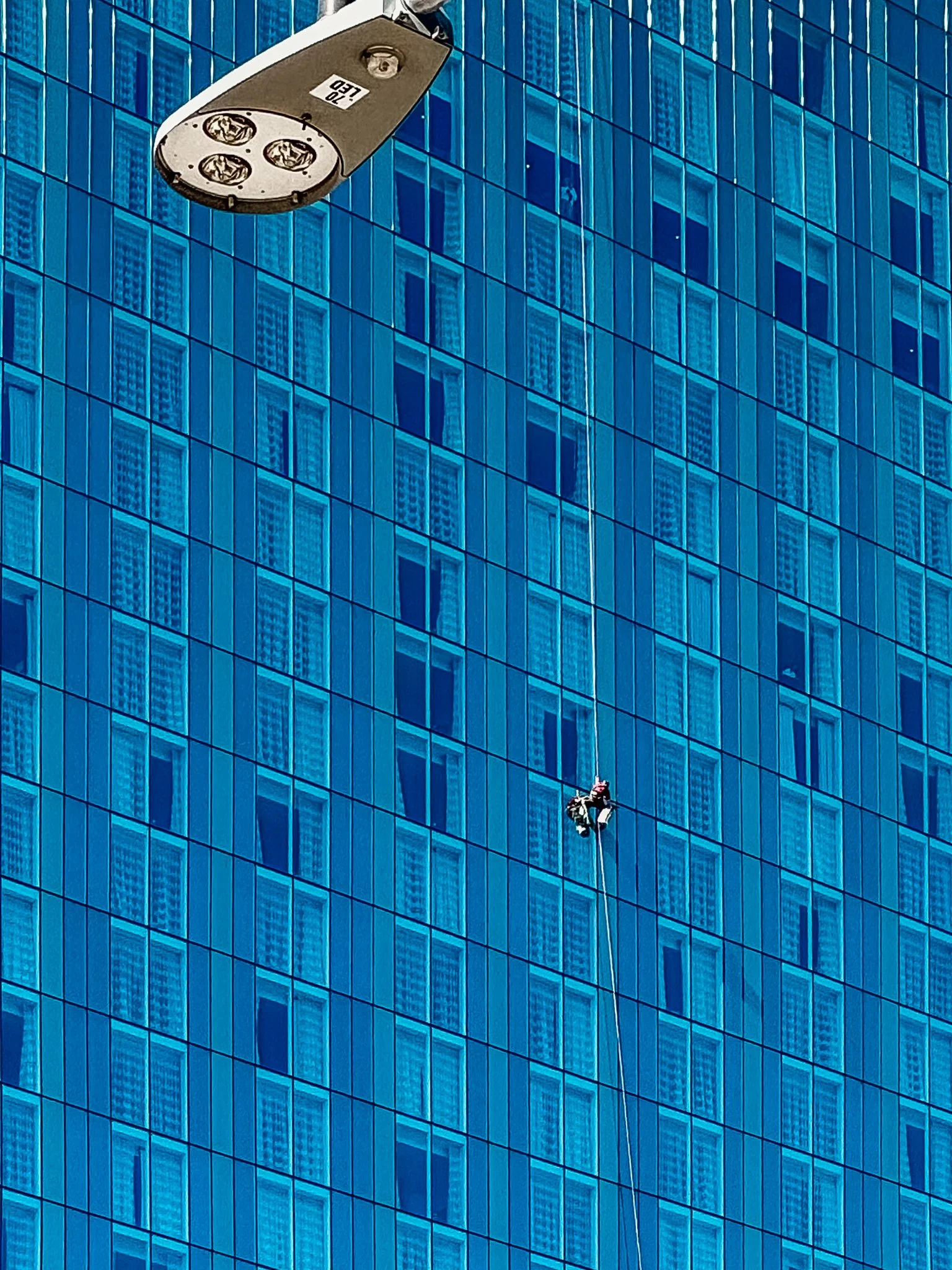 A window washer cleaning a high-rise building with a harness and safety equipment on a rope, against a blue glass facade reflecting nearby buildings.
