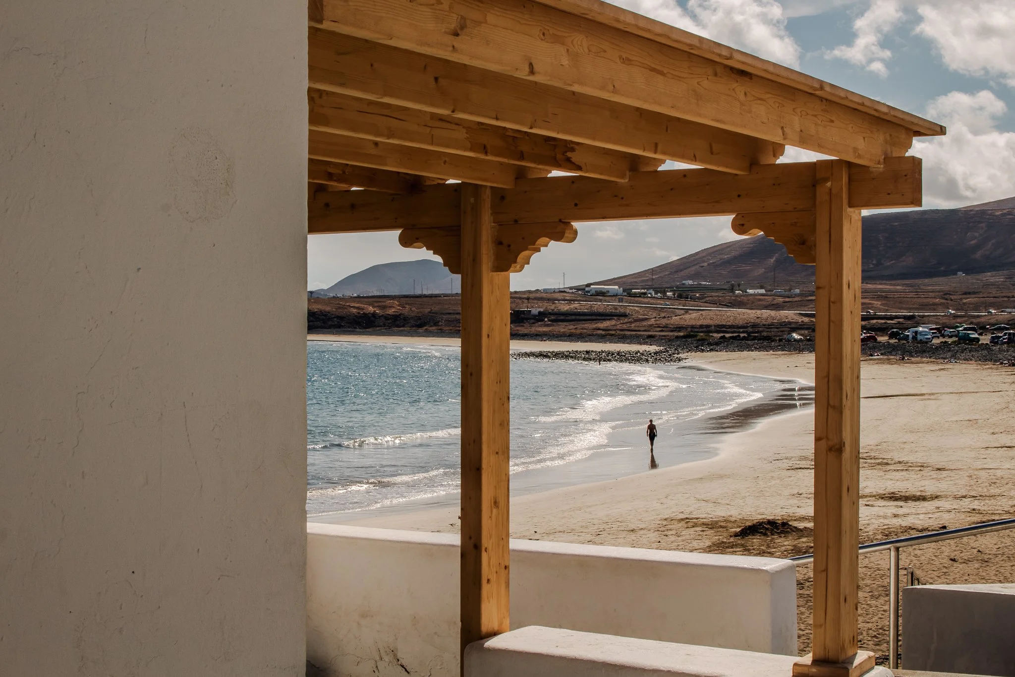 View from a patio overlooking a sandy beach, with a wooden roof structure in the foreground and a person walking along the shoreline, with mountains and a cloudy sky in the background.