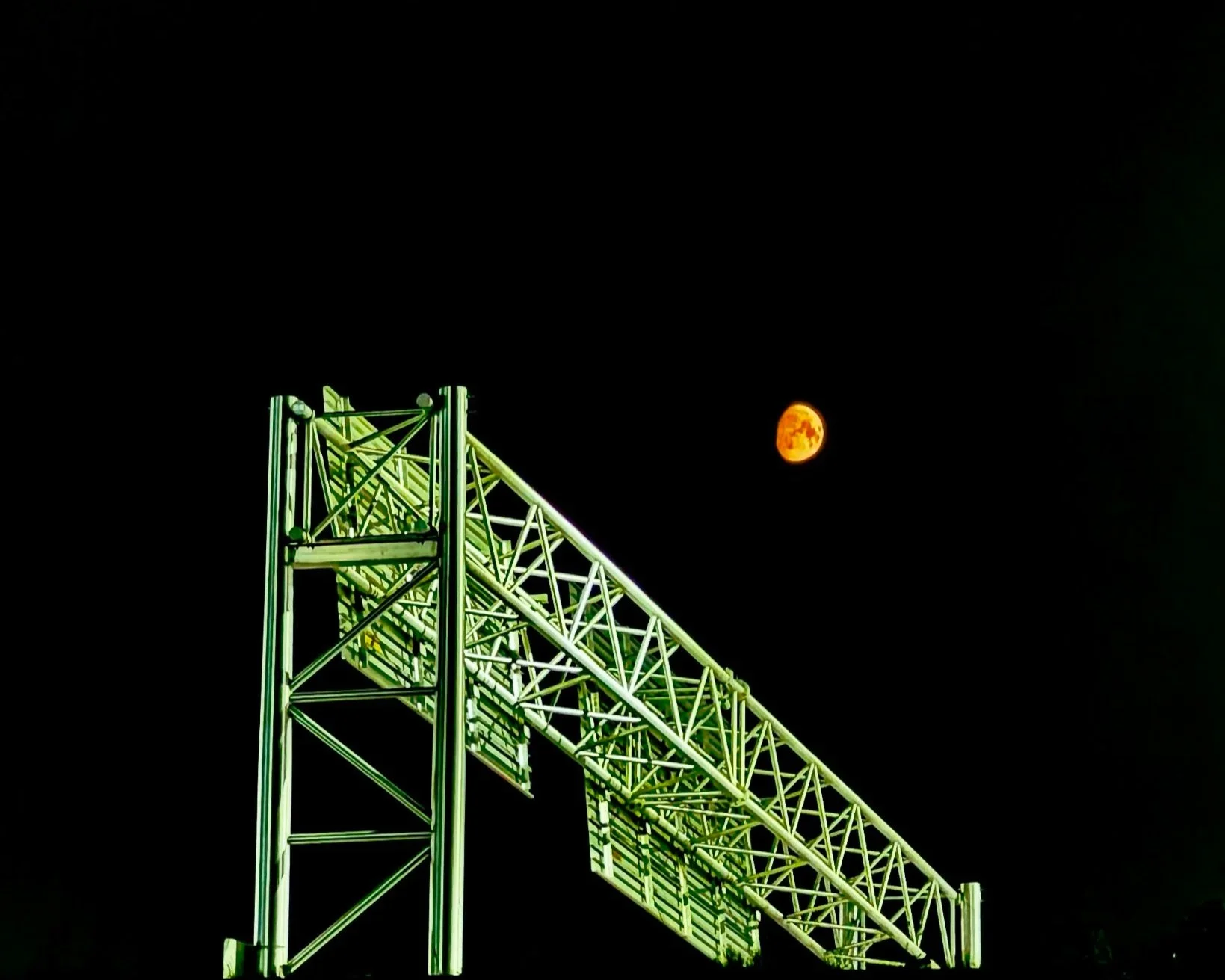 A green metal structure, possibly a highway sign or billboard frame, against a dark night sky with an orange moon in the background.