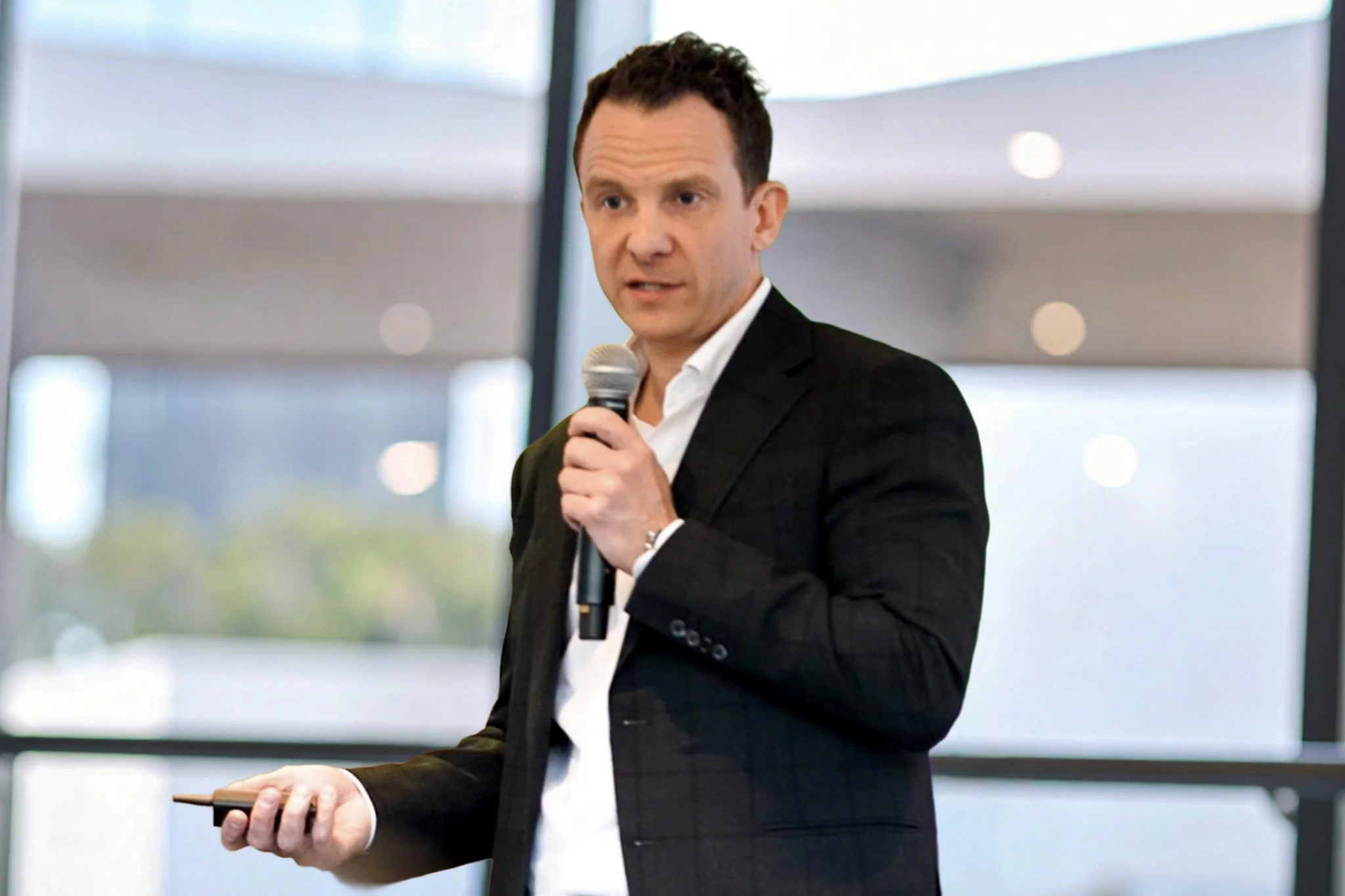 Man in a black suit holding a microphone and a remote, speaking at a conference indoors with large windows in the background.