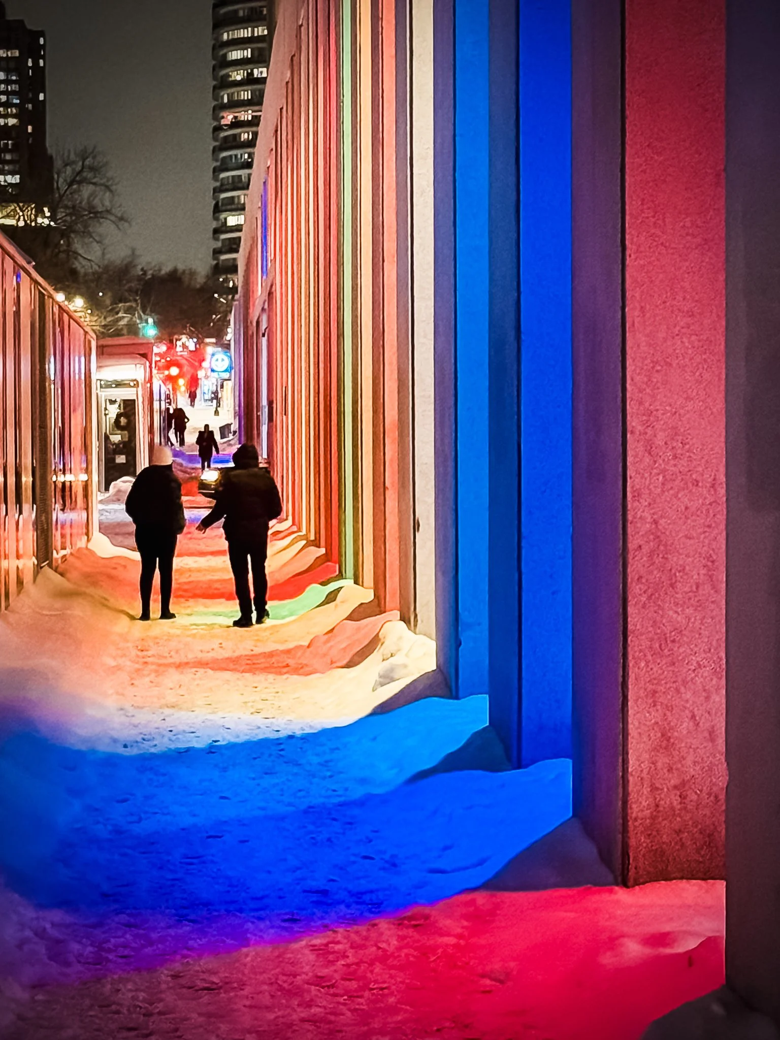 Night view of a snow-covered sidewalk with two people walking, colorful illuminated wall of vertical panels in red, pink, purple, blue, and green, and city buildings in the background.