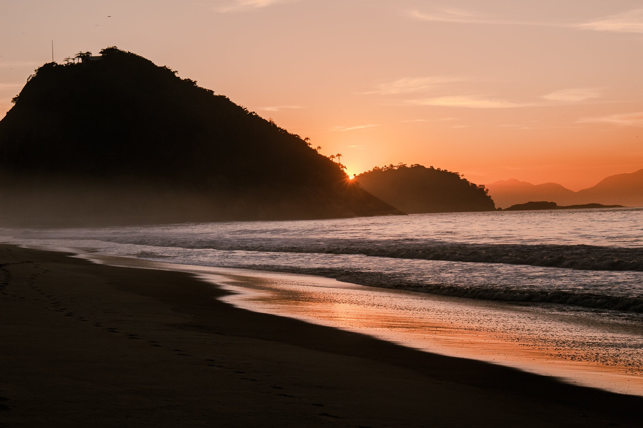 Sunset over a beach with mountains and hills in the background, gentle waves on the shoreline, and footprints in the sand.