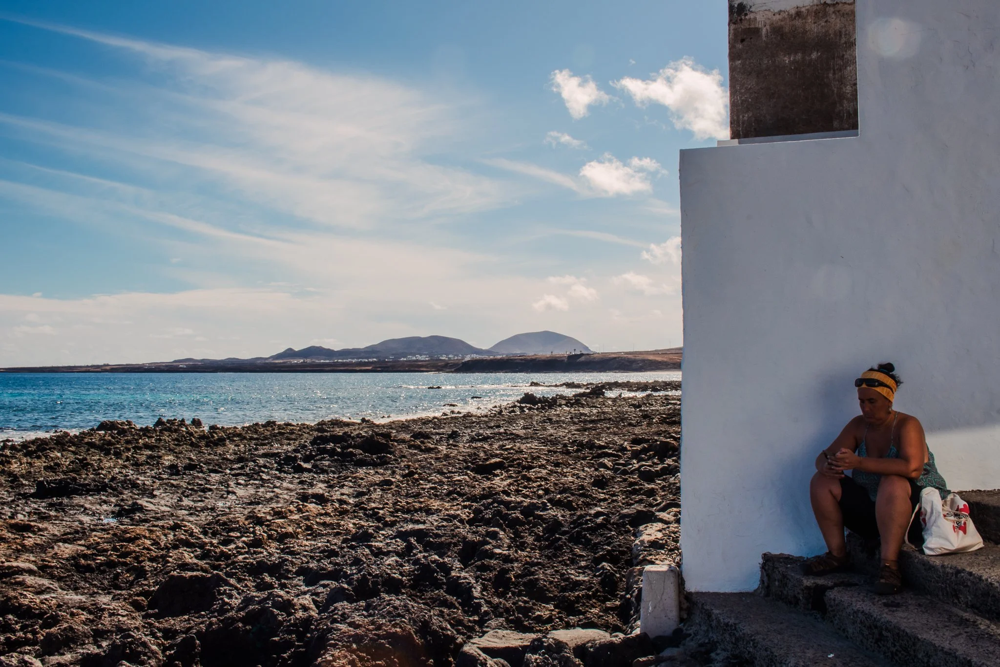 A woman sitting on steps against a white wall by the sea, looking at her phone, with volcanic rocks and an ocean view in the background under a partly cloudy sky.