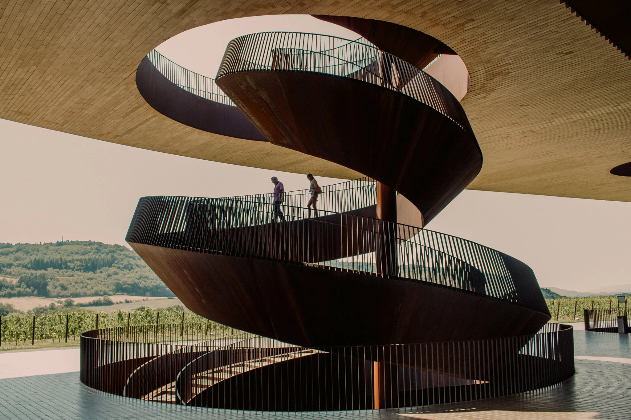 People walking on a spiral staircase inside a modern architectural structure with a large circular opening at the top, overlooking a rural landscape.