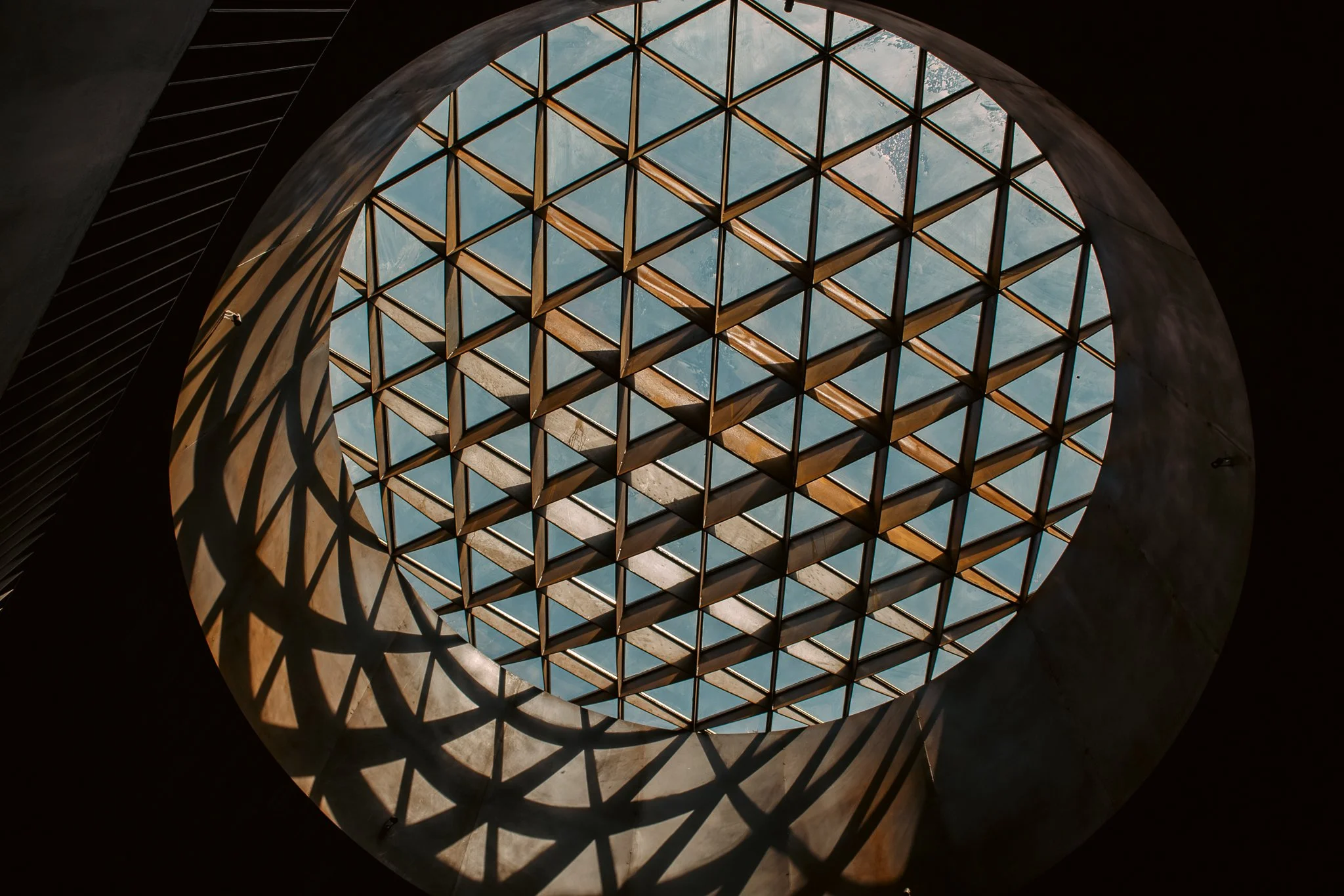 Looking up through a circular skylight with a diamond-patterned grid, casting intricate shadows inside a building.