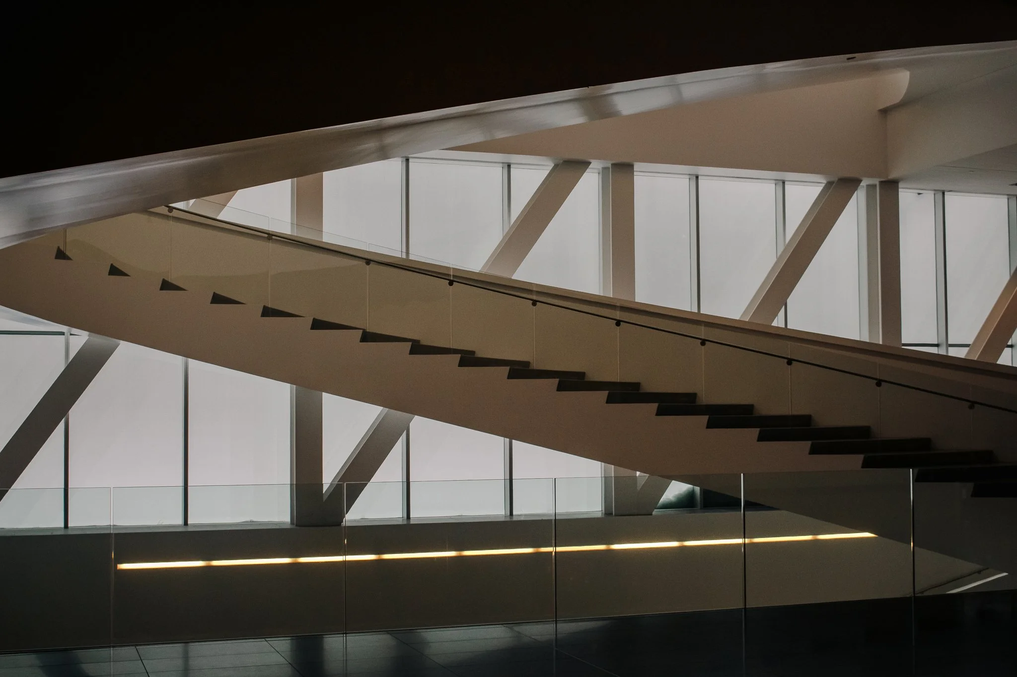 Modern staircase with black steps and glass railing inside a building with large windows and steel support beams.