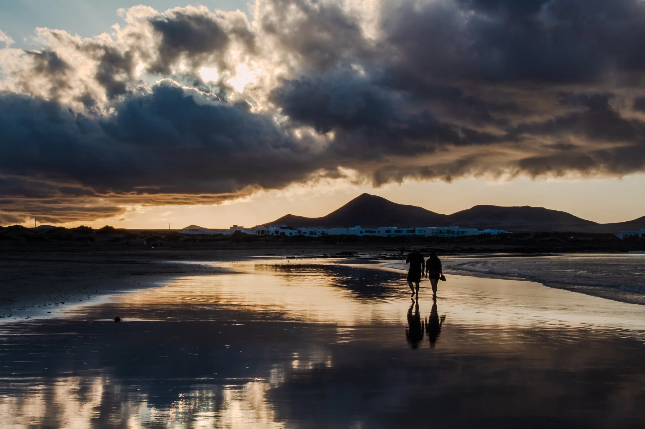 Two people walking along the beach at sunset with dark clouds overhead and mountains in the background, their reflections visible in the wet sand.