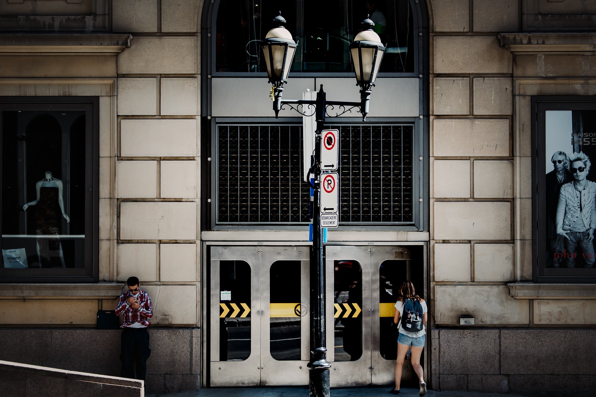 A city street scene with a lamp post in front of a building. Two people are on either side of the lamp post: a man on the left looking at his phone and a woman on the right with a backpack, entering through a door. The building has large windows and 