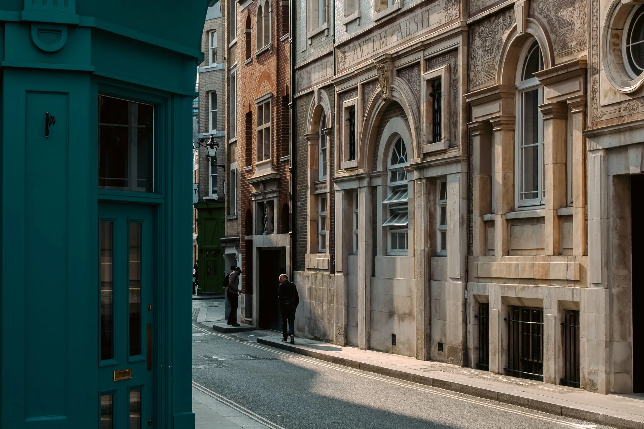 An urban city street scene featuring a teal building on the left and historic brick and stone buildings on the right, two men standing and talking near the sidewalk.