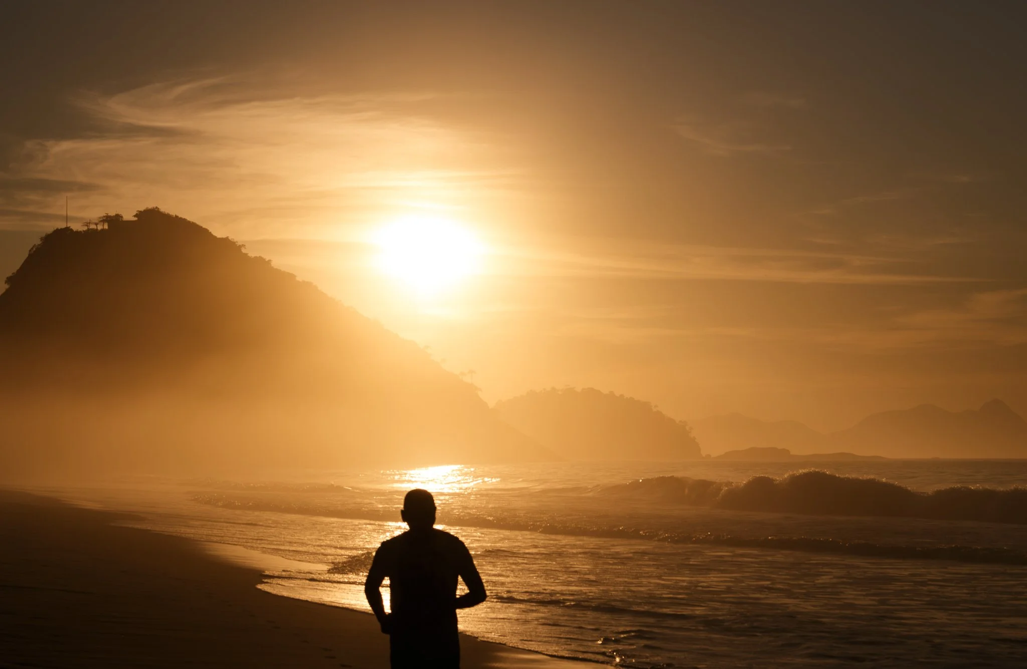 A person jogging along the beach at sunset with the sun setting behind a mountain.