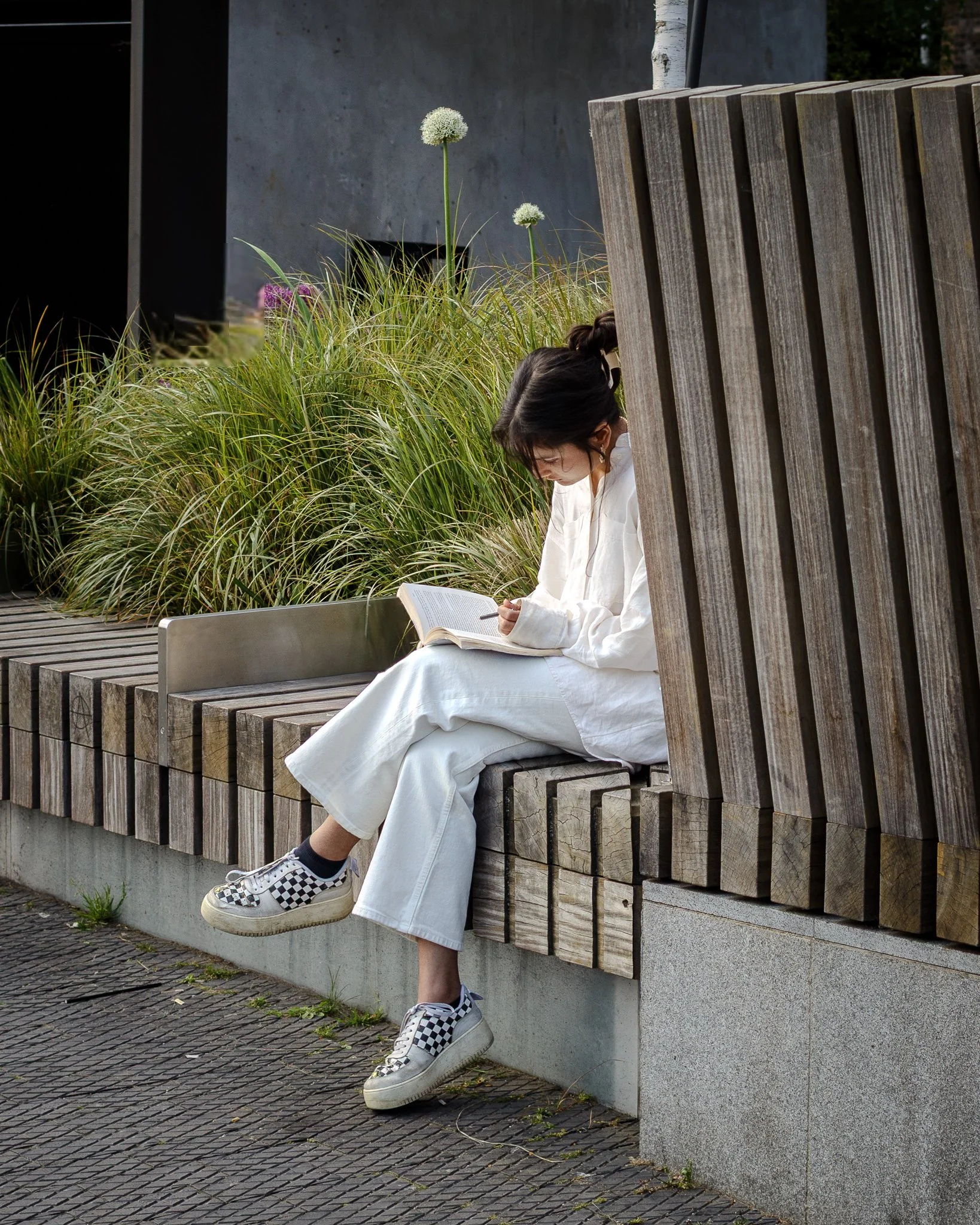 A woman sitting on a wooden bench reading a book outdoors, surrounded by green plants and flowers.