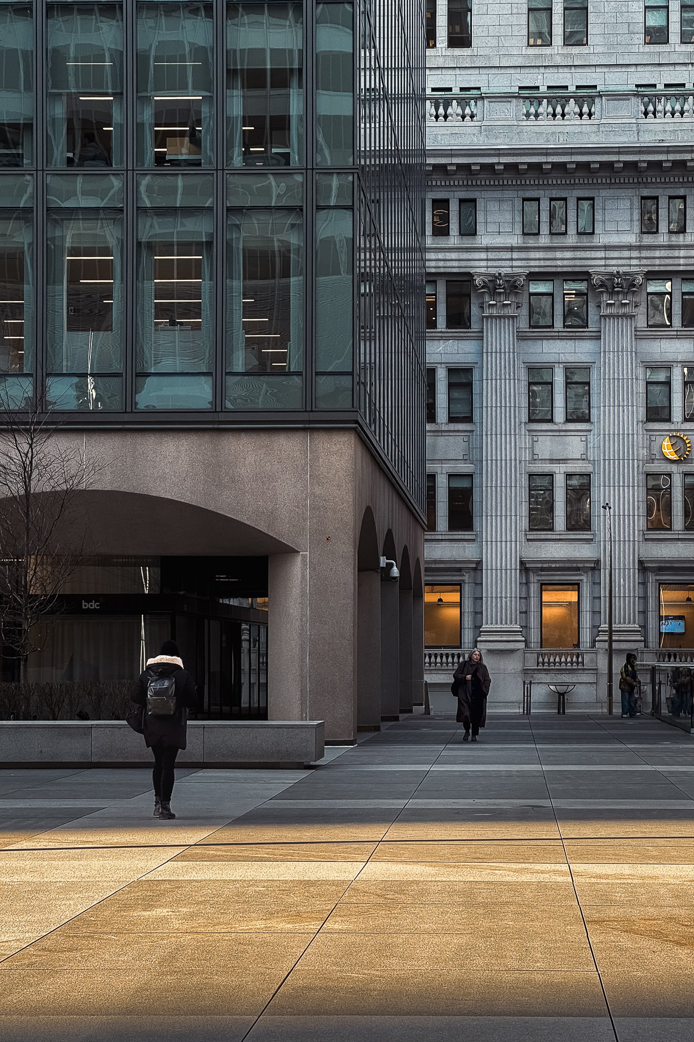 Urban street scene with two pedestrians walking past modern glass and traditional stone buildings.