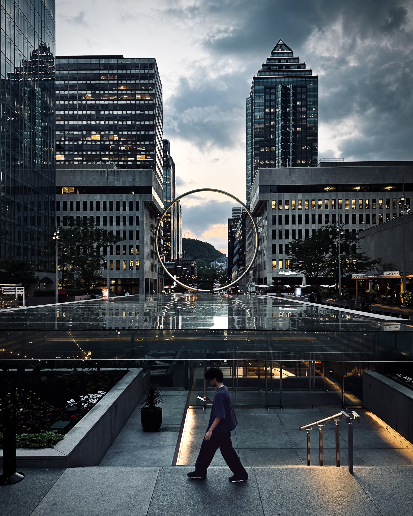 A cityscape with tall modern glass office buildings, a large illuminated circular sculpture, and a person walking on the sidewalk looking at their phone at dusk.