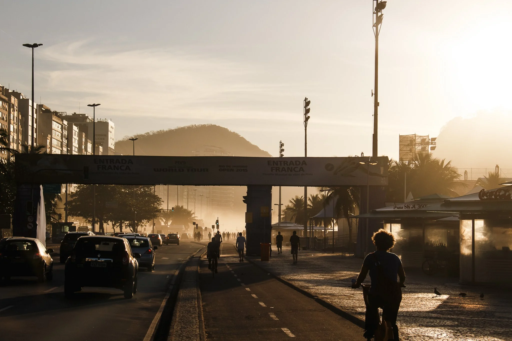People walking and biking on a city street at sunset with buildings, trees, and a bridge.