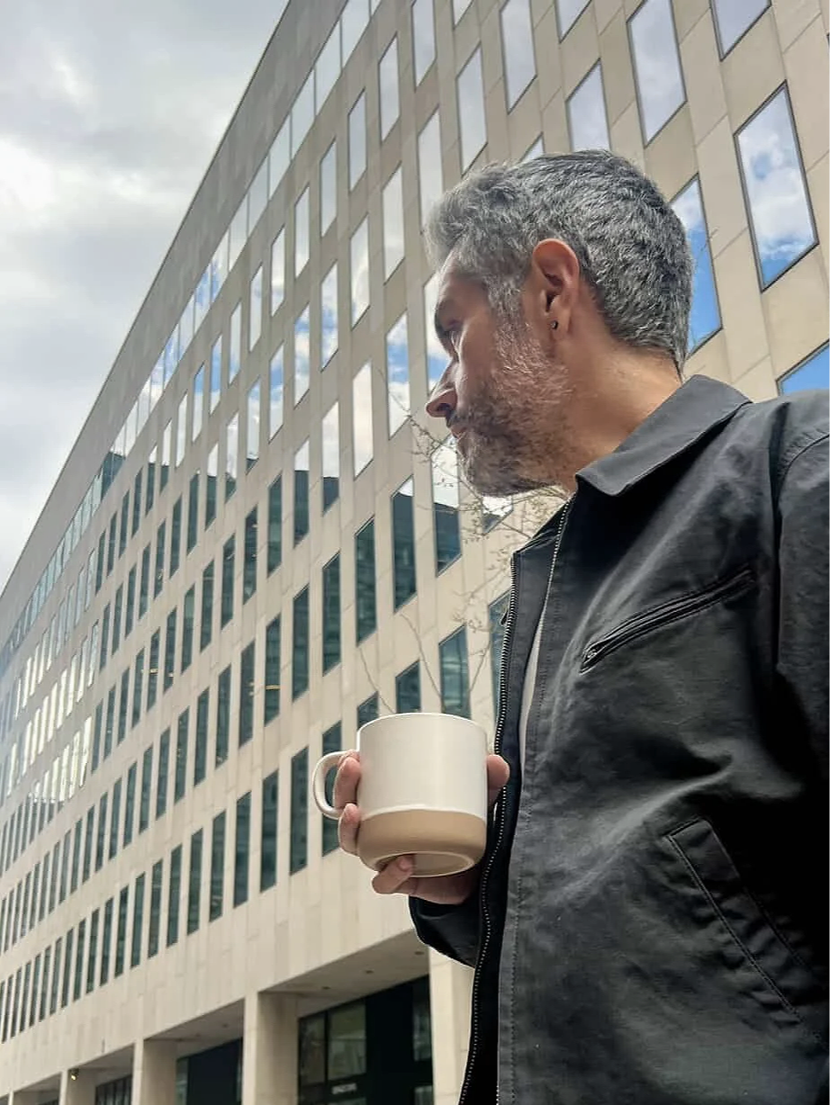 A man with gray hair and beard holding a beige coffee mug outdoors, standing near a tall modern office building with large windows and a partly cloudy sky in the background.