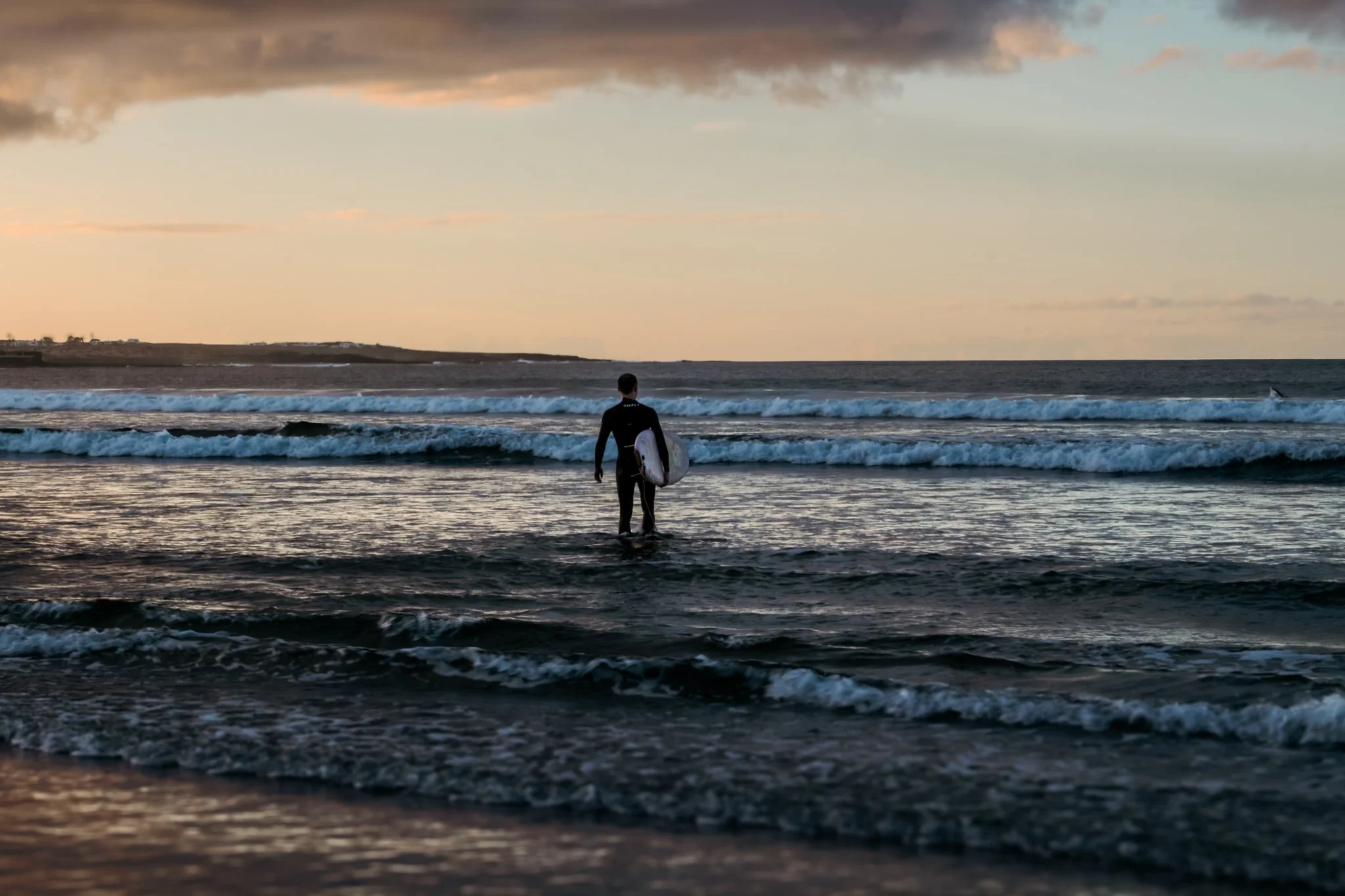 A surfer walking into the ocean at sunset holding a surfboard, with waves and a cloudy sky in the background.