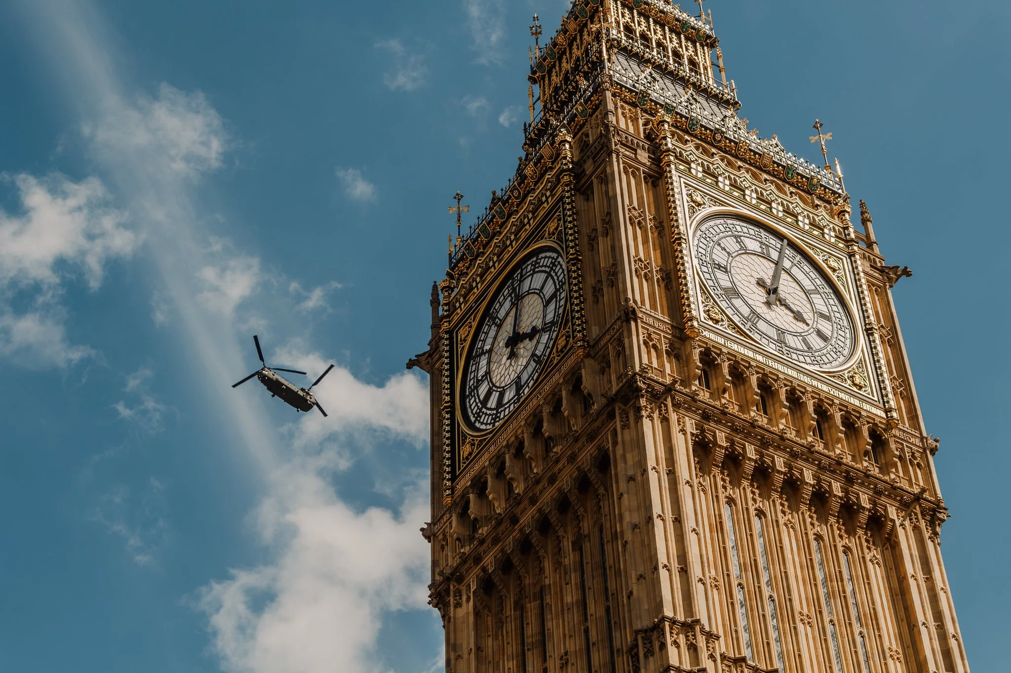 Close-up of the Elizabeth Tower, commonly known as Big Ben, with a helicopter flying nearby against a partly cloudy blue sky.