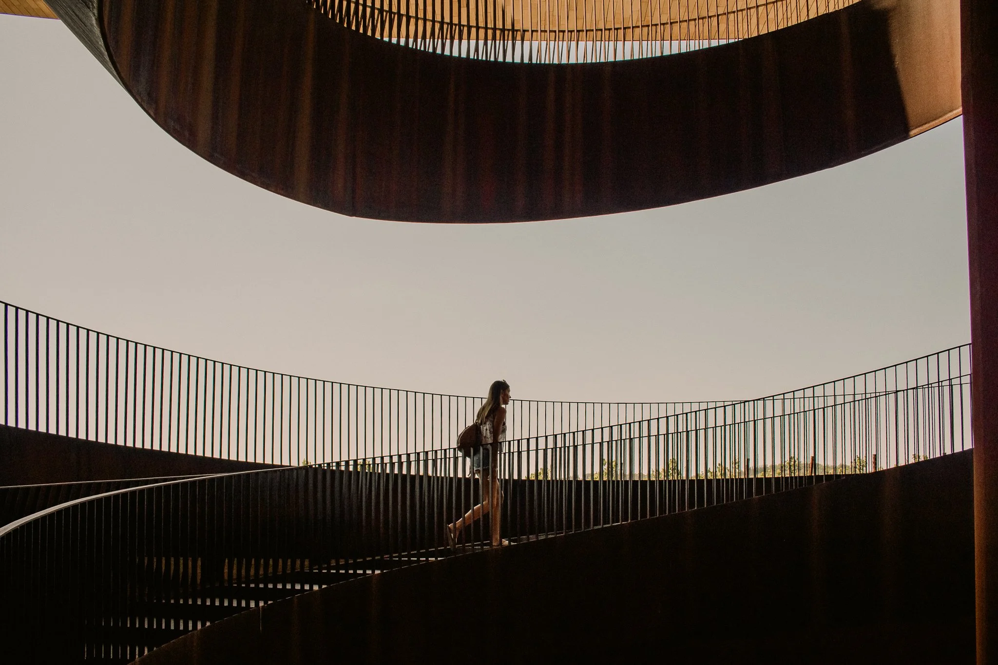A woman with a backpack walking on a curved outdoor staircase with metal railings, with a large circular structure overhead and a clear sky background.