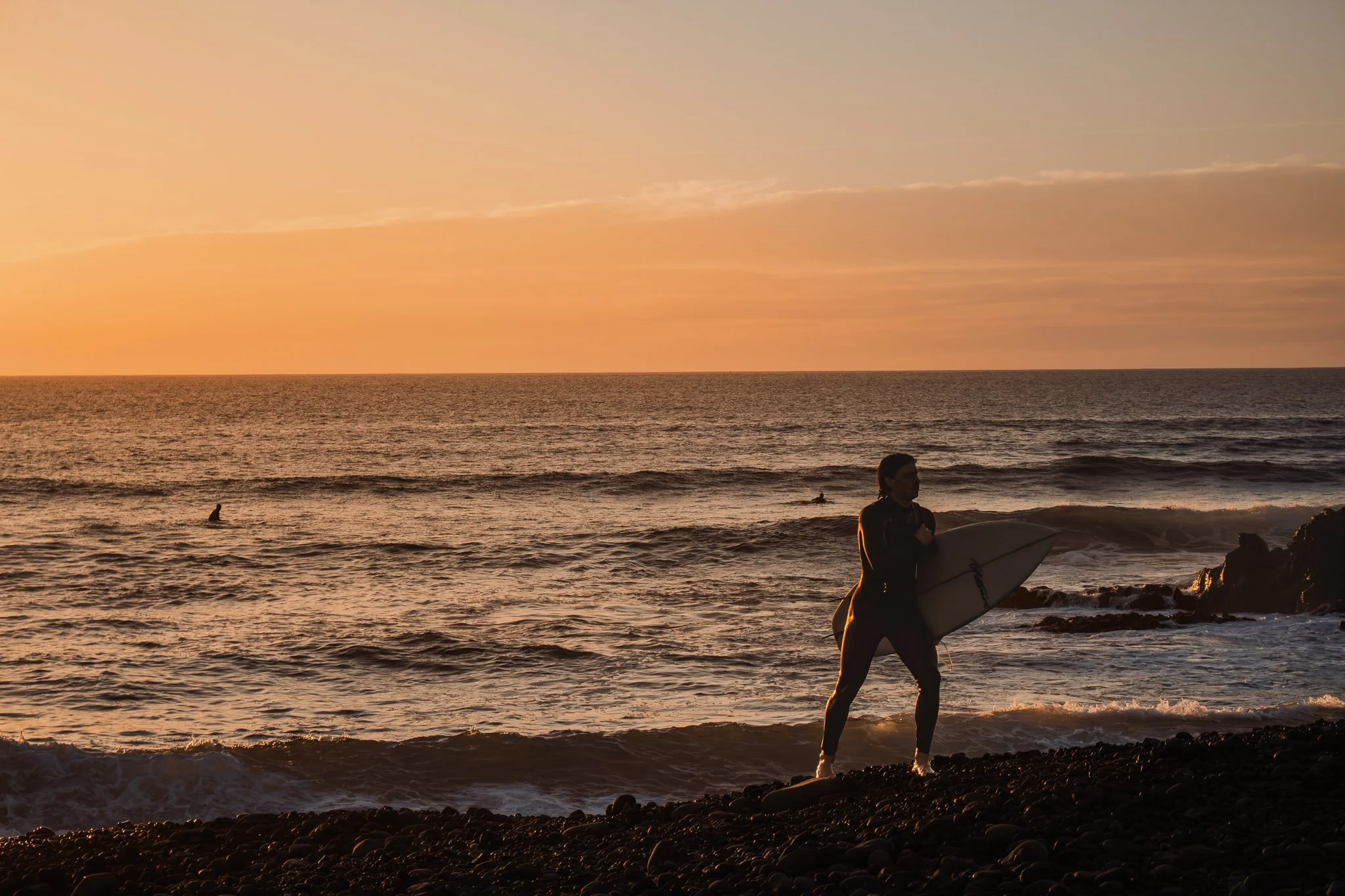 A person in wetsuit holding a surfboard standing on a rocky beach at sunset, with the ocean and other surfers in the background.