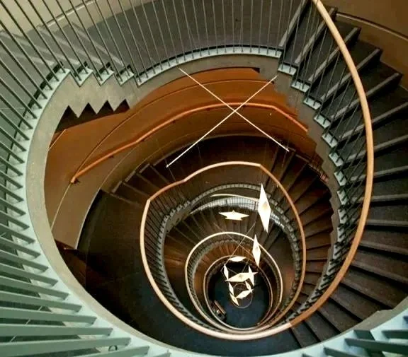 Looking down the center of a spiral staircase with dark steps and light-colored handrails, creating a visual vortex effect.