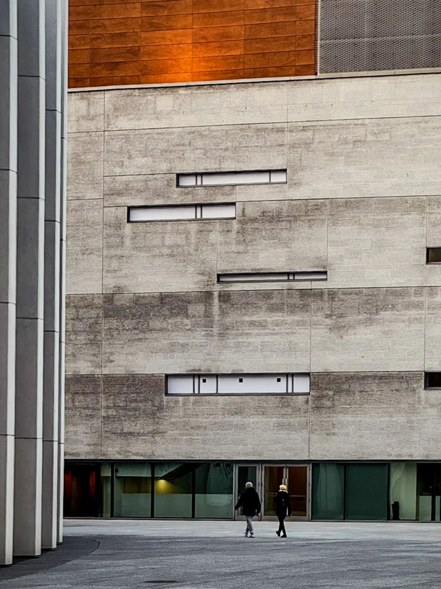 Two people walking in front of a modern building with concrete and glass exterior, and narrow horizontal windows.