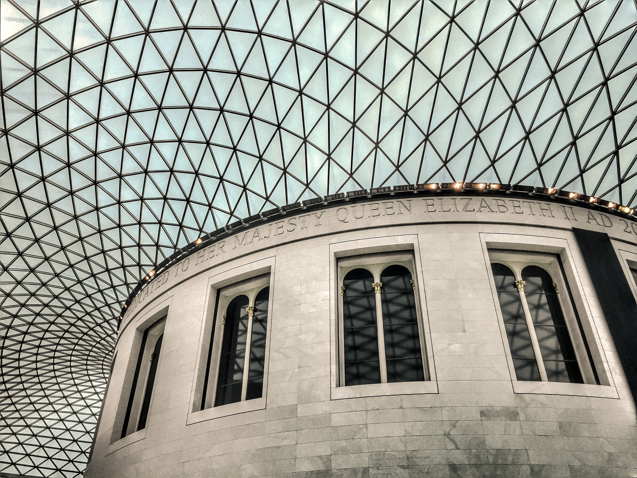 Interior of a building featuring a curved stone wall with windows and an intricate glass ceiling with a geometric pattern.