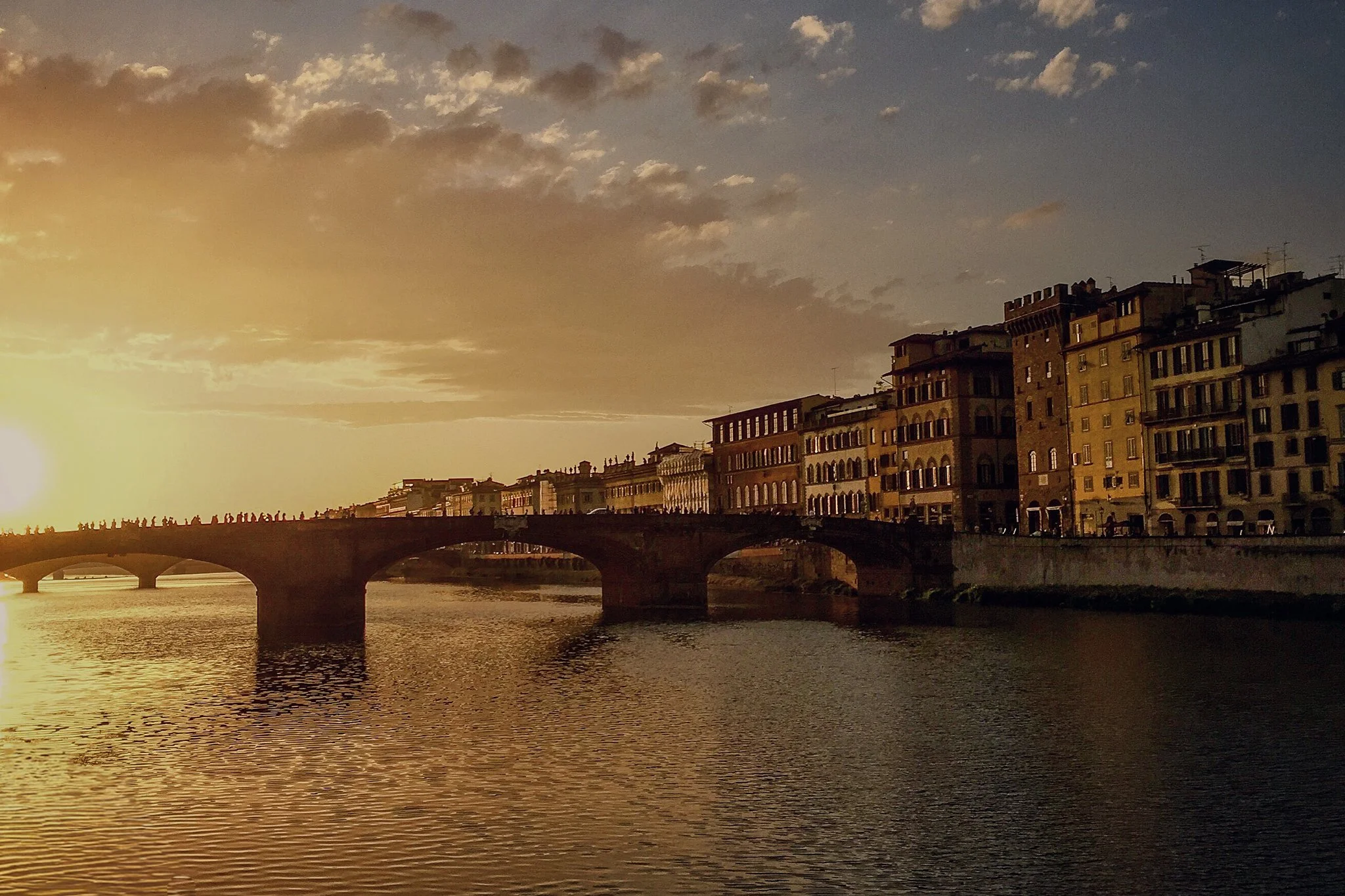Sunset over a river with a stone bridge and colorful buildings along the waterfront in Florence, Italy.