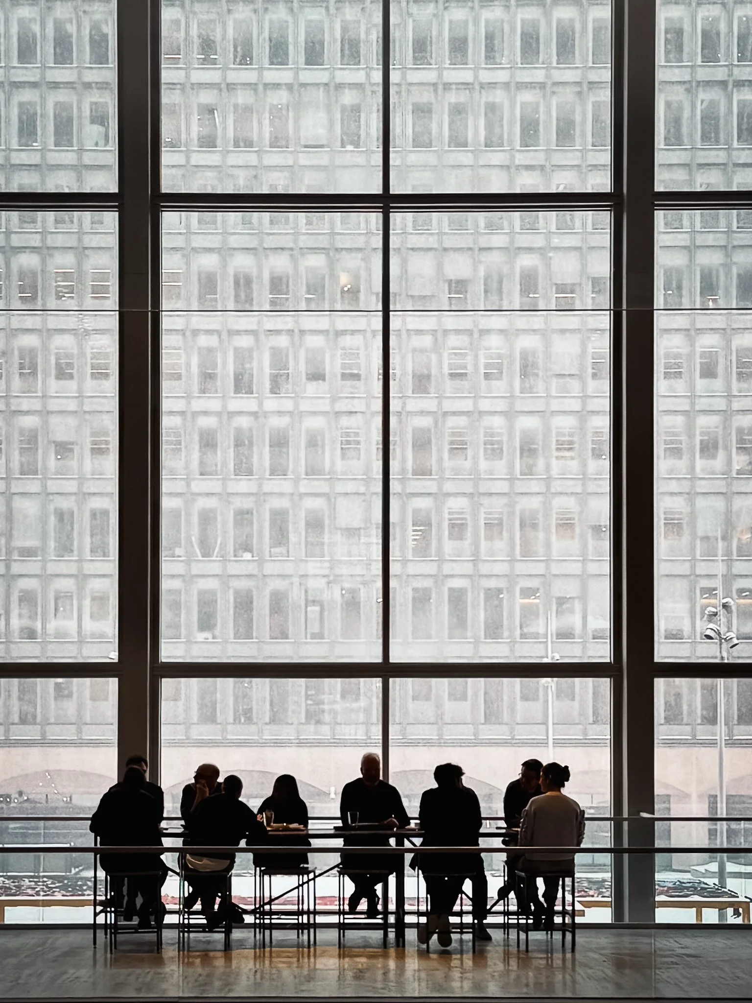Silhouettes of people sitting at a table in front of large windows with a city building facade in the background.
