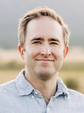 Portrait of a smiling man outdoors with a blurred landscape in the background.