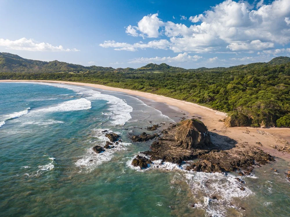 Aerial view of a sandy beach with rocks, ocean waves, lush green forest, and mountains in the background under a partly cloudy sky.
