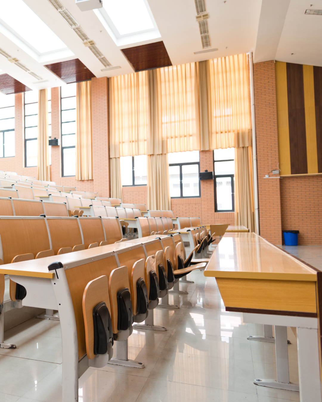 Empty lecture hall with wooden seats, large windows with beige curtains, and a wooden desk at the front.